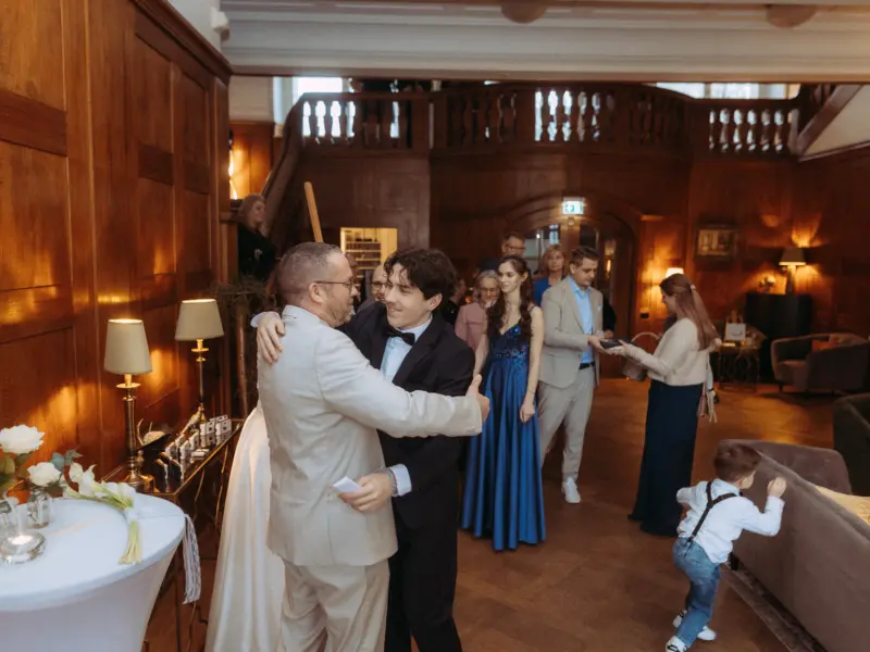 Ein herzlicher Moment bei einer Hochzeit: Ein Bräutigam im schwarzen Anzug umarmt einen Gast in einem hellen Sakko in einer eleganten Hotellobby. Im Hintergrund sind weitere Hochzeitsgäste und ein spielendes Kind zu sehen – festgehalten vom Hochzeitsfotograf in Schwerin und MV.
