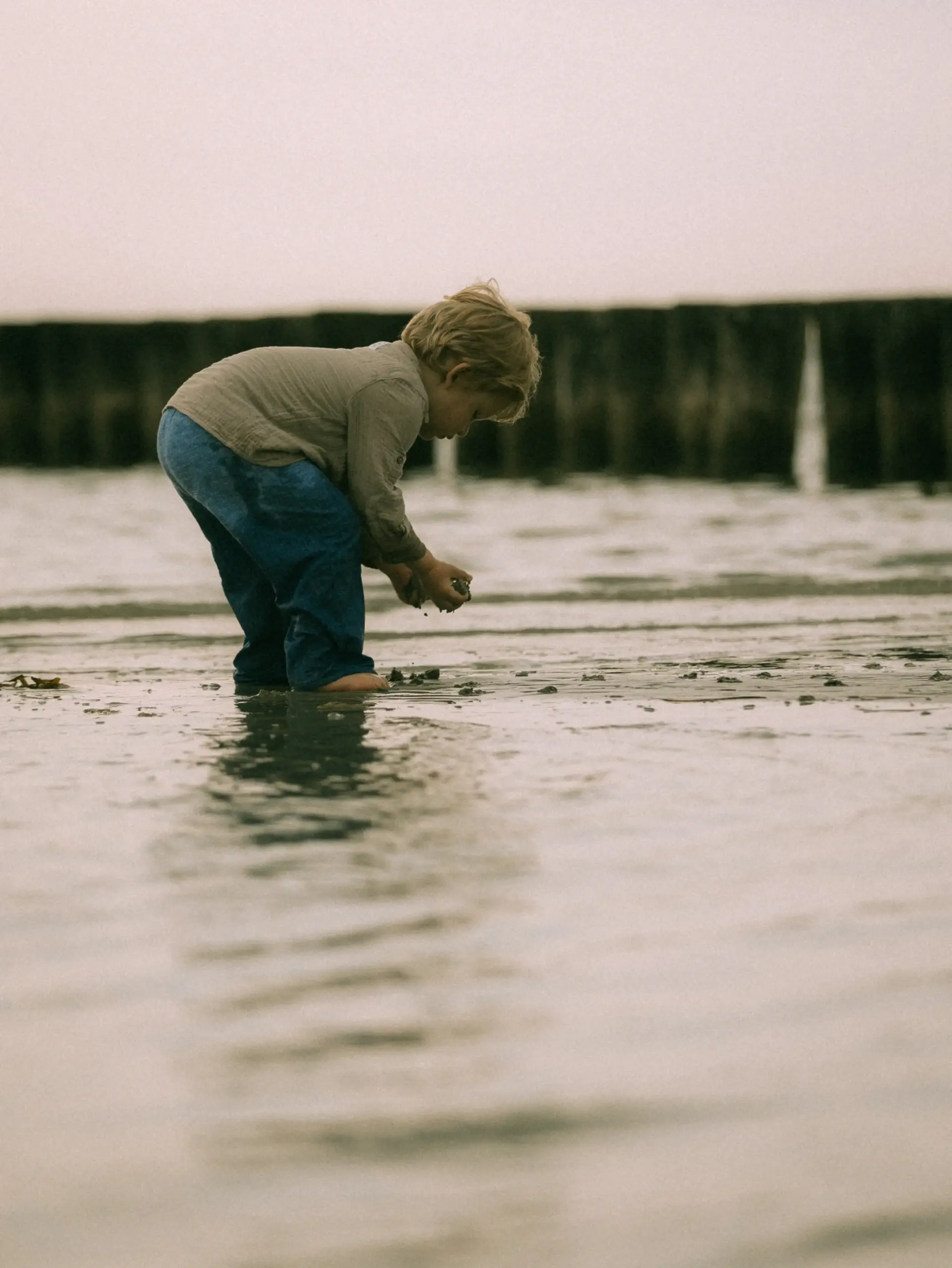 Ein kleines Kind mit blonden Haaren spielt vertieft im flachen Wasser am Strand. Es trägt ein beiges Hemd und eine blaue Hose, während im Hintergrund hölzerne Pfähle im Meer zu sehen sind. Ein stimmungsvoller Moment für Familienfotos in Schwerin.