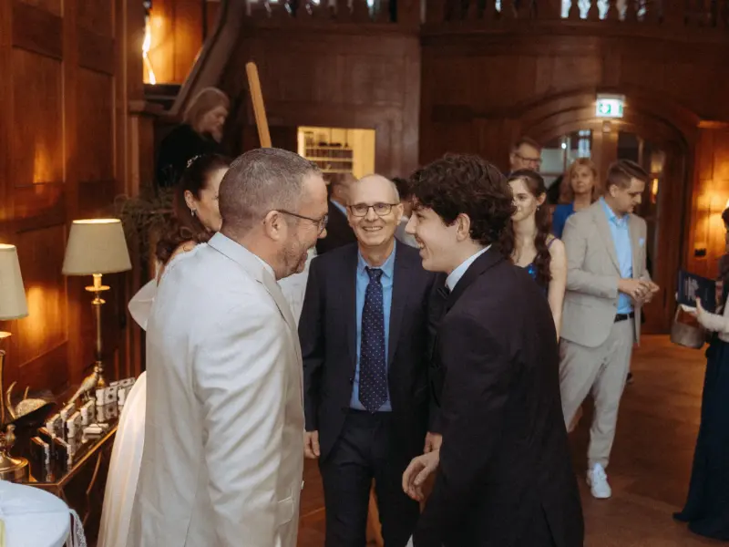 Ein herzliches Gespräch zwischen zwei Männern in Anzügen während einer Hochzeitsfeier in einem eleganten, holzgetäfelten Saal. Im Hintergrund lächelt ein Mann in einem blauen Anzug, während weitere Gäste in festlicher Kleidung den Raum beleben. Hochzeitsfotograf in Schwerin und MV.