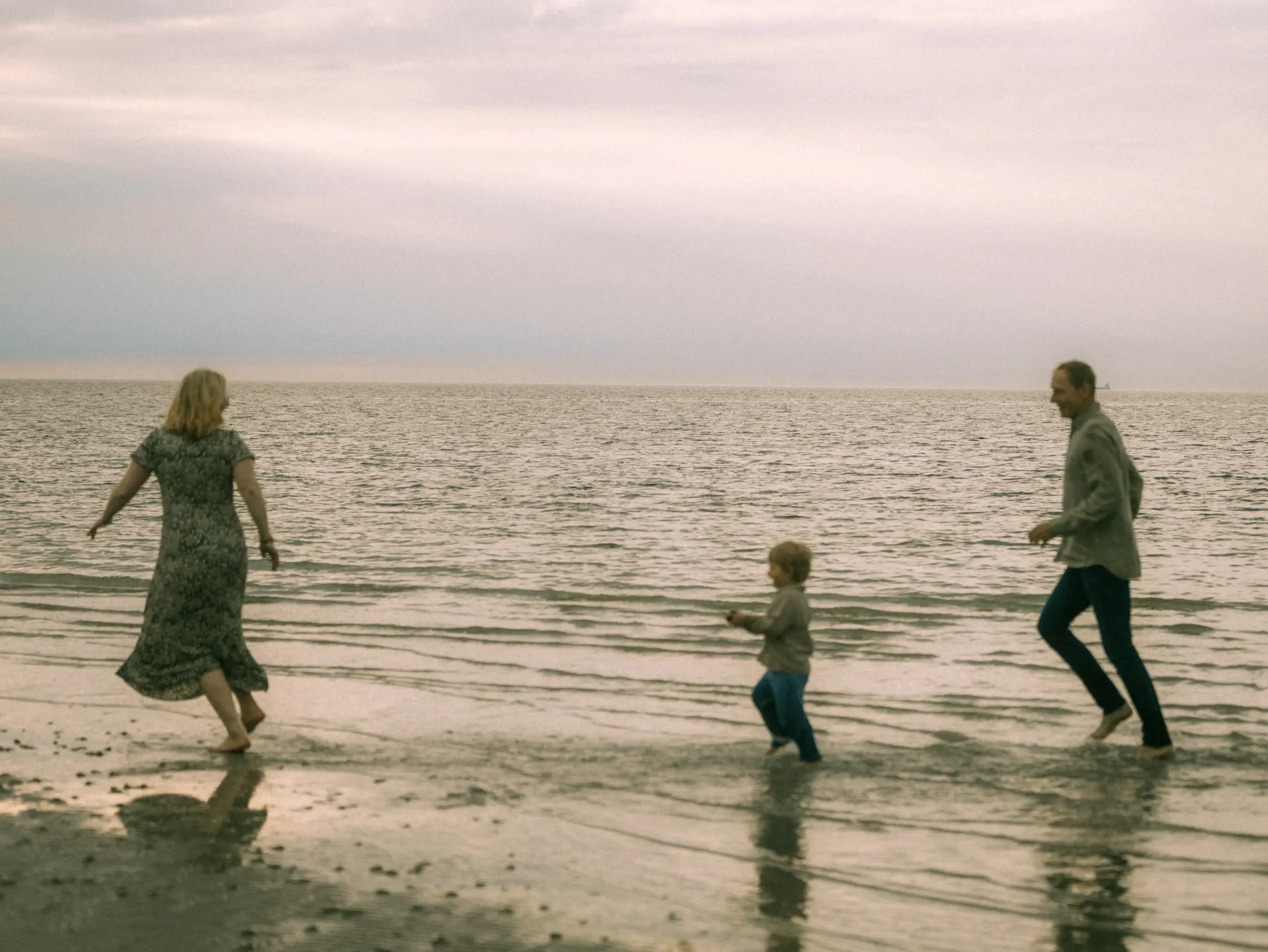 Eine glückliche Familie rennt barfuß durch das flache Wasser am Strand bei Sonnenuntergang. Die Eltern und ihr Kind genießen die gemeinsame Zeit am Meer, eingefangen von einem professionellen Familienfotograf in Schwerin bei einem entspannten Fotoshooting in natürlicher Atmosphäre.