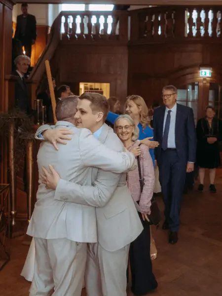 Zwei Männer in hellgrauen Anzügen umarmen sich herzlich bei einer Hochzeitsfeier in einem eleganten, holzgetäfelten Raum. Im Hintergrund lächeln Gäste, während ein Hochzeitsfotograf in Schwerin und MV diesen emotionalen Moment der Verbundenheit einfängt.