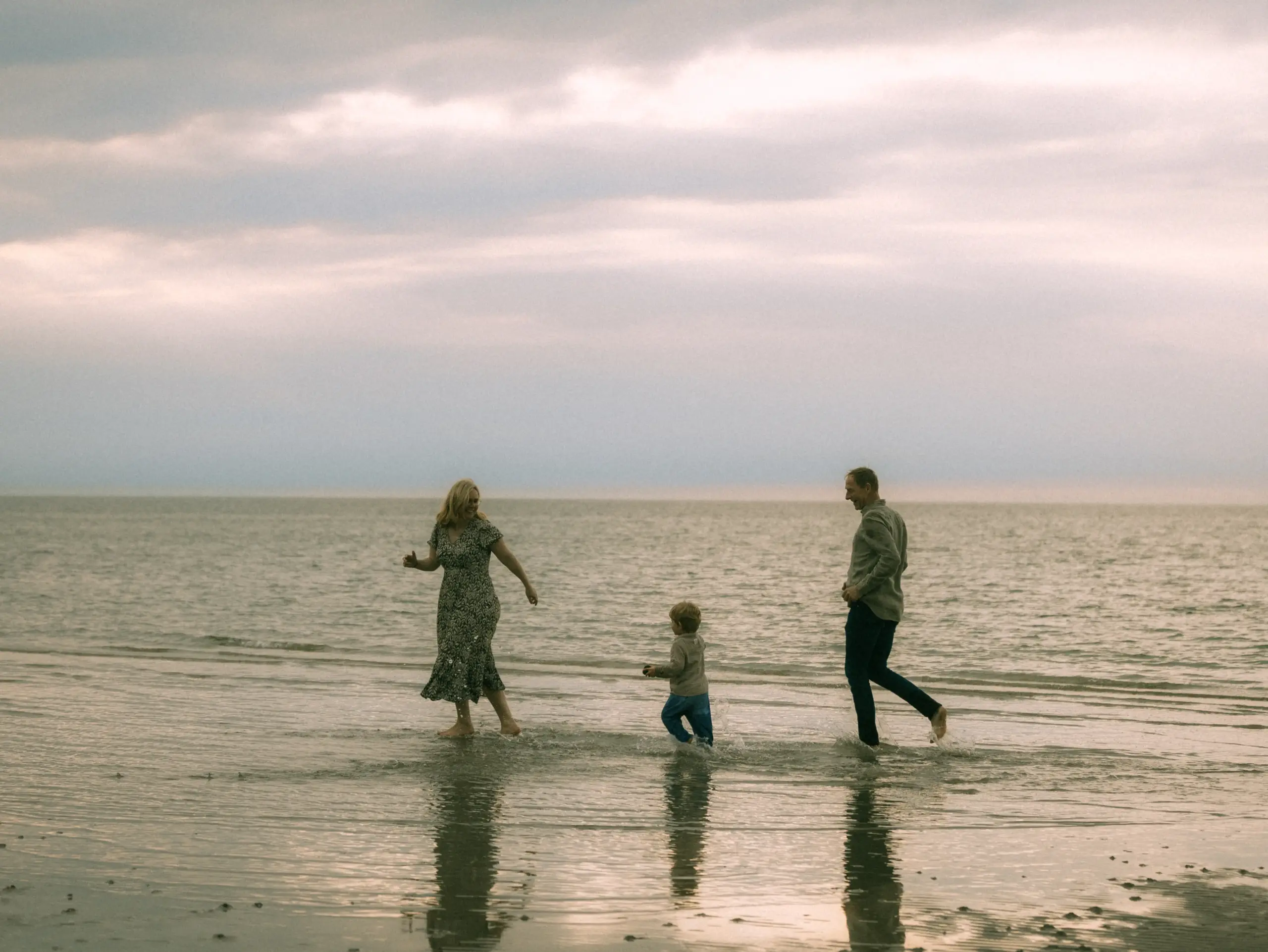 Eine junge Familie genießt einen glücklichen Moment am Strand, während sie gemeinsam durch das flache Wasser laufen. Die Eltern und ihr kleines Kind strahlen Lebensfreude aus, eingefangen von einem professionellen Familienfotograf in Schwerin bei einem entspannten Shooting am Meer.
