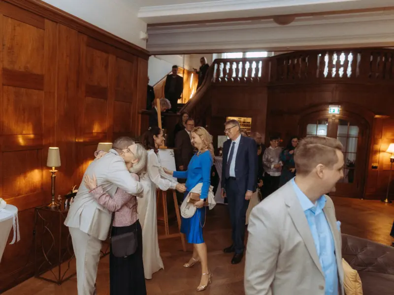 Ein herzlicher Empfang bei einer festlichen Feier: Ein Paar umarmt sich zur Begrüßung, während Gäste im Hintergrund in einem eleganten, holzvertäfelten Saal stehen. Professionelle Momente festgehalten von einem Hochzeitsfotograf in Schwerin und MV.