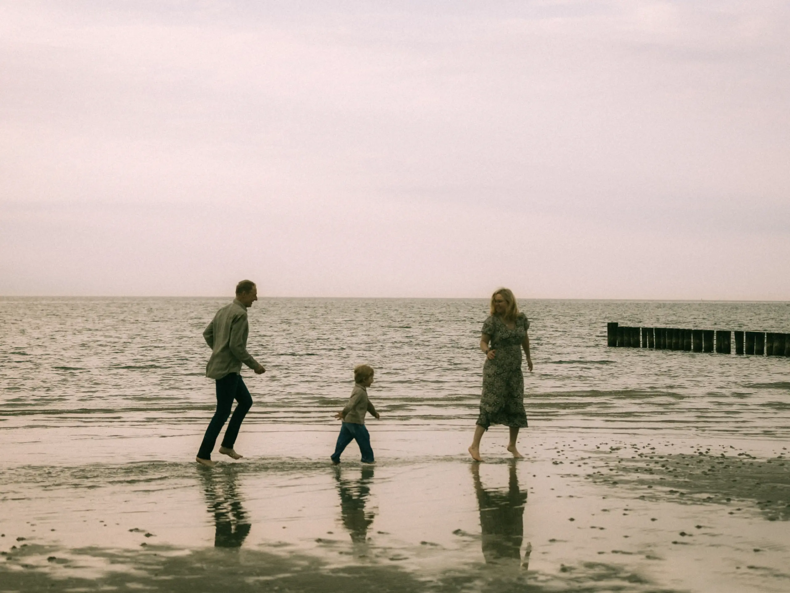 Eine junge Familie rennt lachend und barfuß durch das flache Wasser am Strand bei bewölktem Himmel. Die Eltern und ihr kleiner Sohn genießen einen entspannten Moment am Meer. Authentische Familienfotos in Schwerin, die eine natürliche und fröhliche Stimmung einfangen.