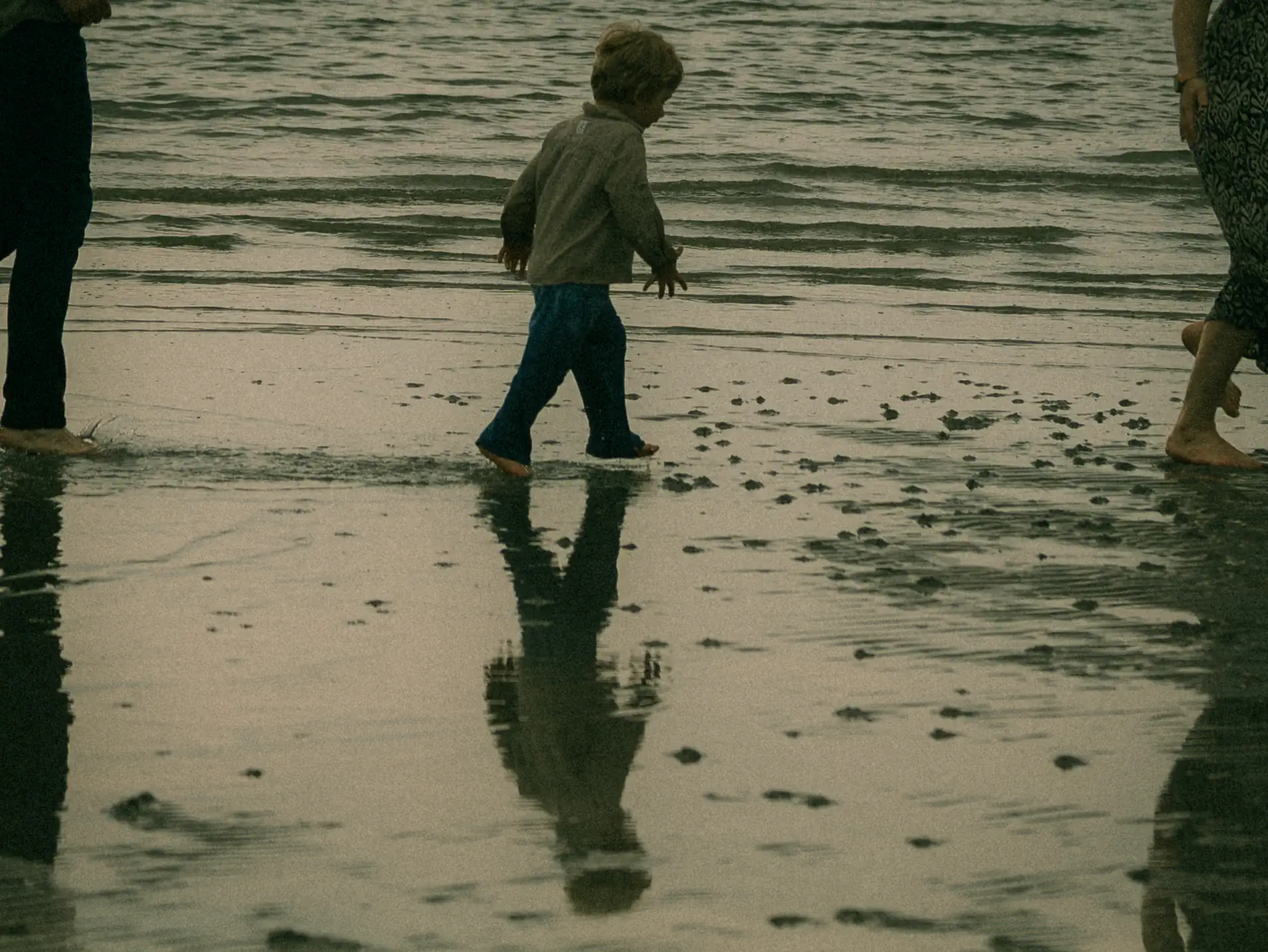 Ein kleines Kind läuft barfuß durch das flache Wasser an einem Strand, während sich seine Silhouette im nassen Sand spiegelt. Begleitet wird es von zwei Erwachsenen, was diesen Moment zu einer wunderbaren Erinnerung macht. Fotograf für Familien in Schwerin.