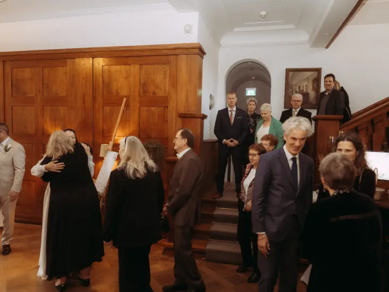 Gäste begrüßen sich herzlich in einem eleganten, holzgetäfelten Raum während einer Hochzeitsfeier. Im Hintergrund stehen weitere Gäste auf einer Treppe, während ein Hochzeitsfotograf in Schwerin und MV die festliche Stimmung und das herzliche Miteinander der Hochzeitsgesellschaft festhält.