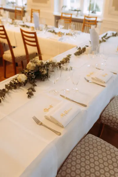 Festlich gedeckte Hochzeitstafel mit weißer Tischdecke, eleganter Blumendekoration aus Eukalyptus und weißen Rosen, goldene Namensschilder auf Servietten sowie Weingläser. Professionell festgehalten von Ihrem Hochzeitsfotograf in Schwerin und MV für eine stilvolle Hochzeitsfeier.