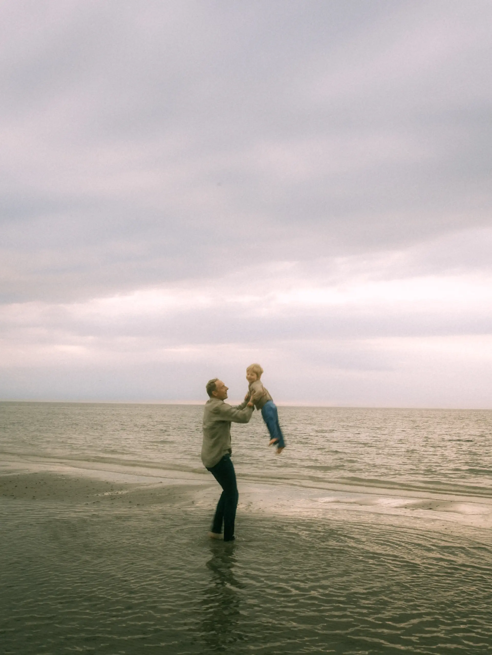 Ein Vater spielt mit seinem kleinen Kind am Strand und hebt es freudig in die Luft, während sie gemeinsam einen Moment voller Glück am Meer genießen. Eine schöne Momentaufnahme für Familienfotos in Schwerin.