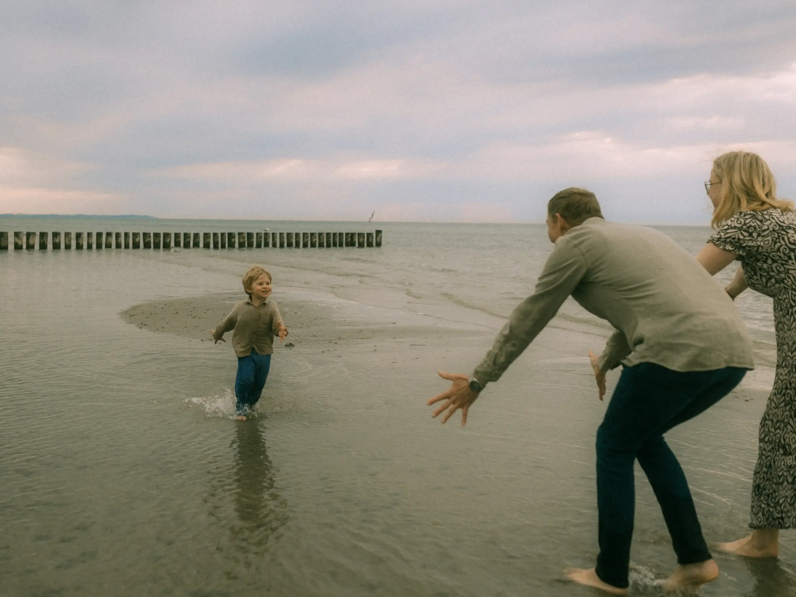 Ein kleiner Junge rennt lachend durch das flache Wasser am Strand auf seine Eltern zu, die ihre Arme ausbreiten, um ihn zu empfangen. Diese herzliche Szene zeigt echte Familienfotos in Schwerin vor der Kulisse des Meeres mit hölzernen Buhnen im Hintergrund.