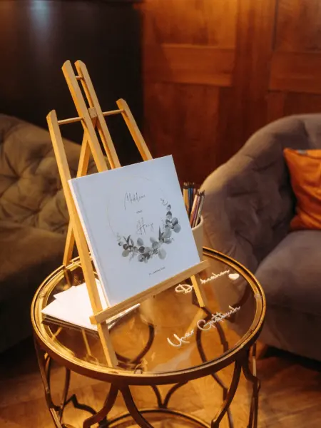Ein elegantes Gästebuch für die Hochzeit von Madeleine und Hajo am 17. Januar 2024, präsentiert auf einer hölzernen Staffelei auf einem goldenen Beistelltisch. Das stimmungsvolle Setting für den Hochzeitsfotograf in Schwerin und MV wird durch Sessel und dezente Dekoration abgerundet.