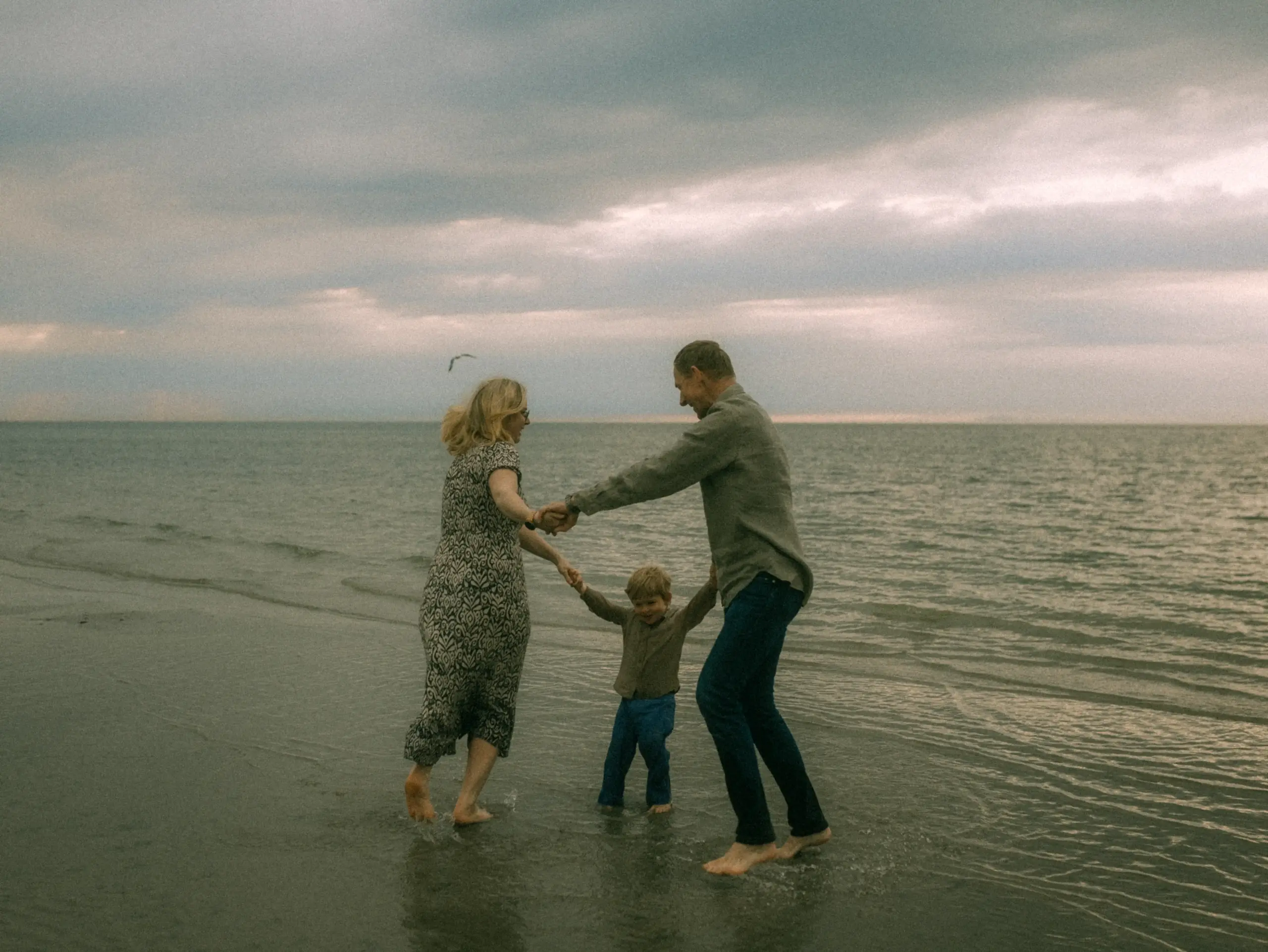 Eine junge Familie genießt einen glücklichen Moment am Strand, während sie sich an den Händen halten und im flachen Wasser tanzen. Die stimmungsvolle Atmosphäre am Meer unter bewölktem Himmel fängt die Freude dieser Familienfotos in Schwerin perfekt ein.