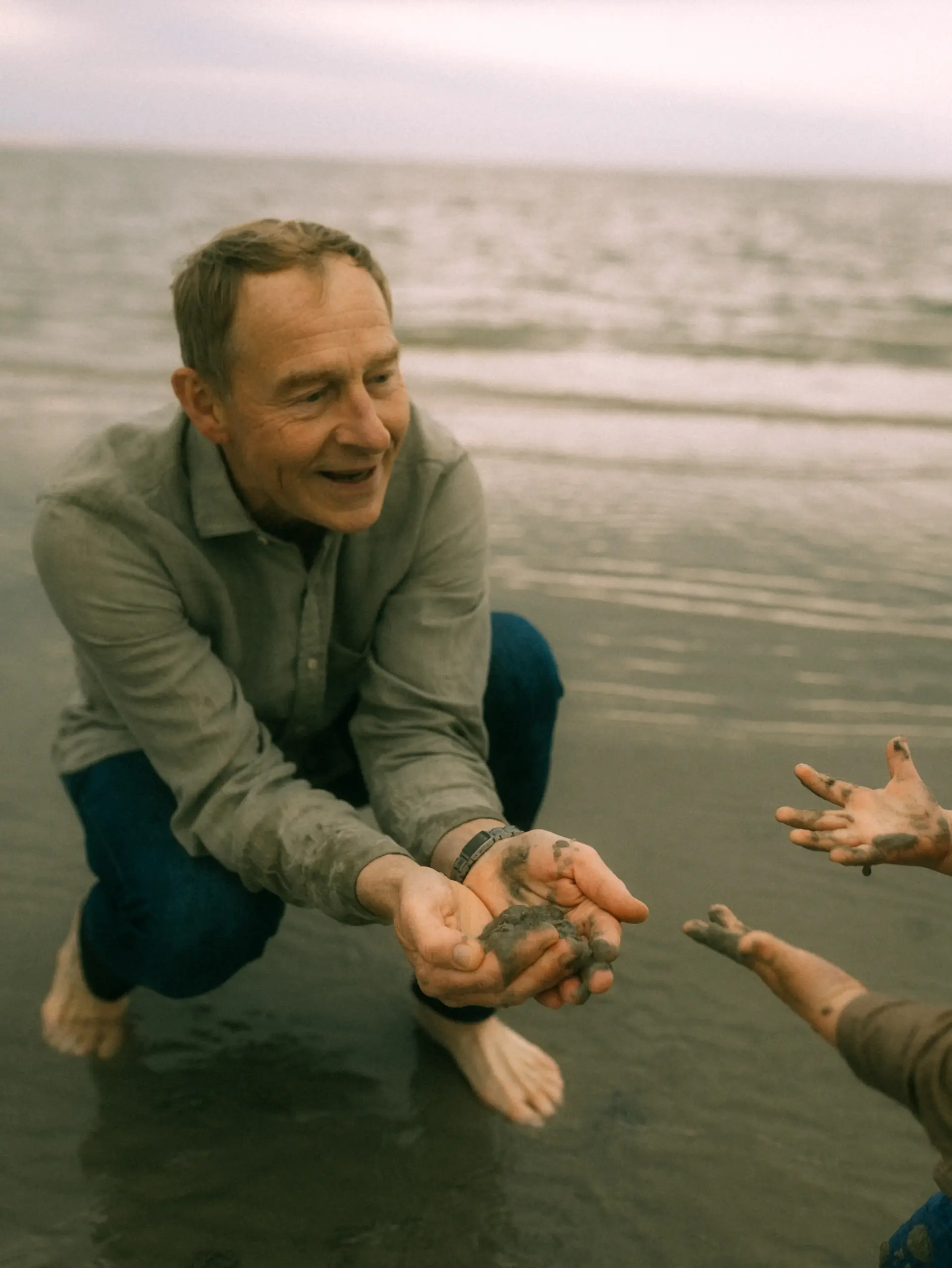 Ein glücklicher Vater kniet am Strand und spielt mit seinem Kind, während sie gemeinsam nassen Sand in den Händen halten. Dieser herzliche Moment wurde von einem professionellen Fotograf für Familien in Schwerin in einer natürlichen Familienfotografie festgehalten.