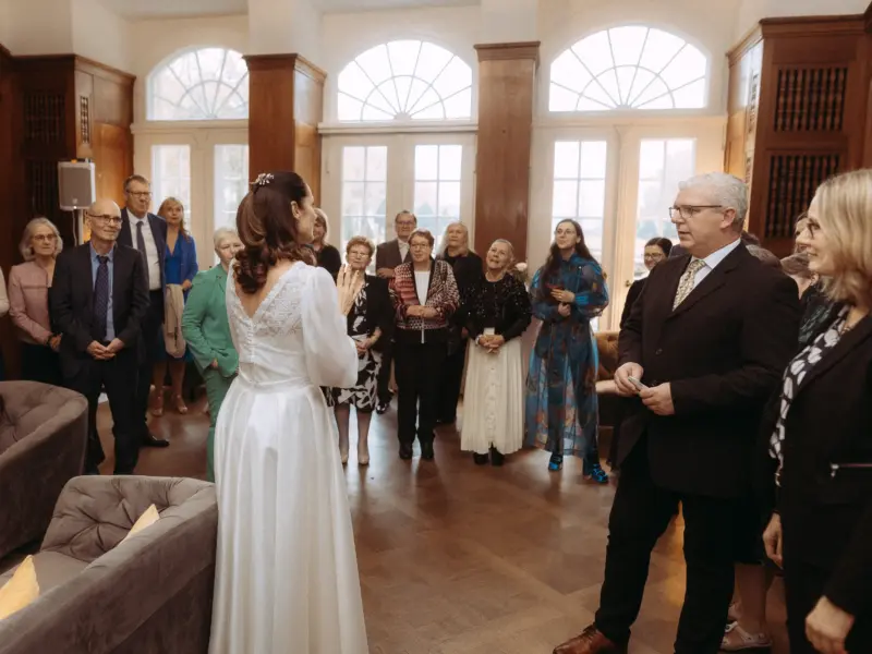 Eine Braut im eleganten weißen Hochzeitskleid spricht zu einer versammelten Hochzeitsgesellschaft in einem hellen, festlichen Saal mit hohen Fenstern. Hochzeitsfotograf in Schwerin und MV, der den emotionalen Moment der Ansprache in einer stilvollen Hochzeitslocation einfängt.