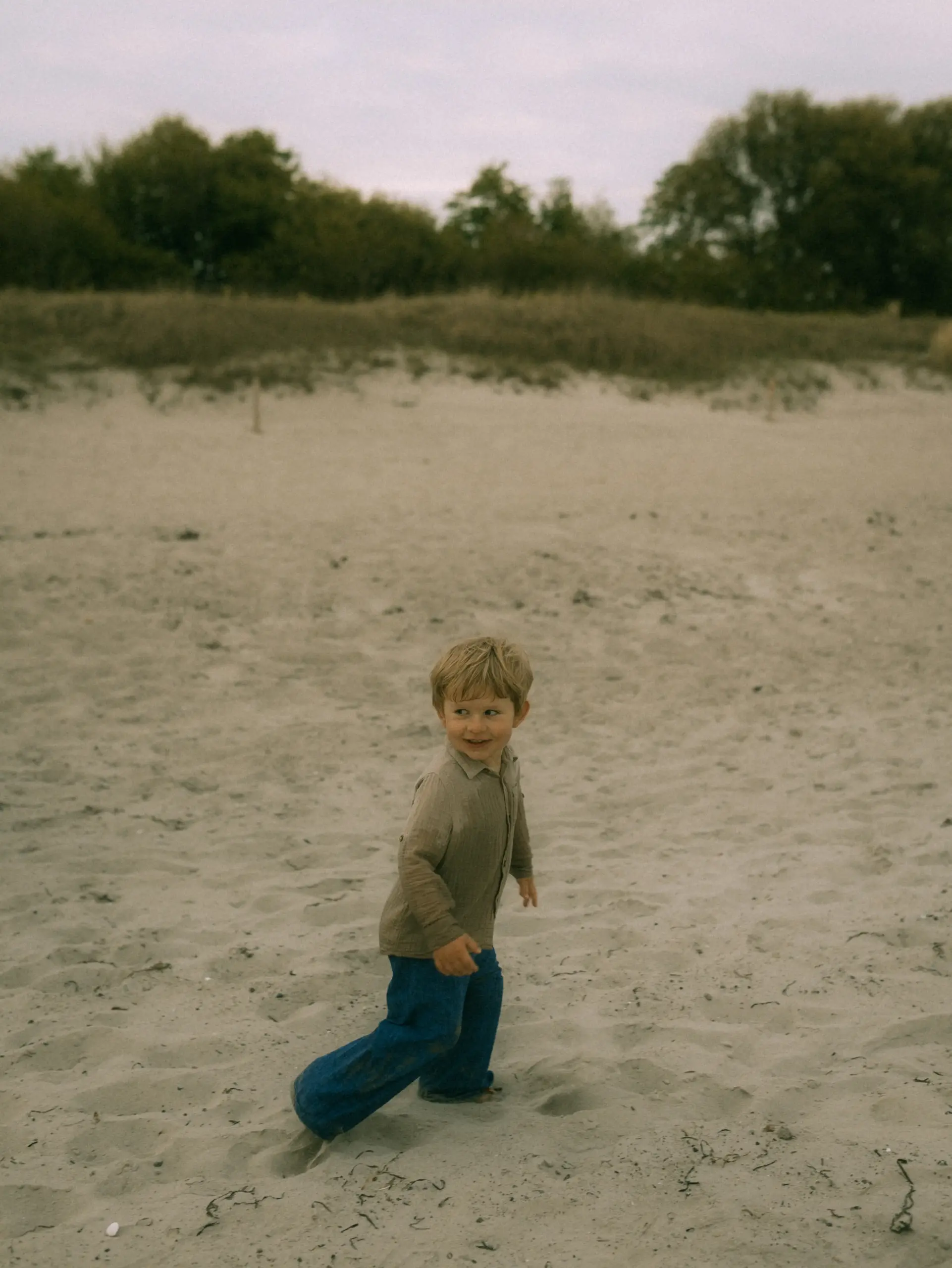 Ein kleines Kind mit blondem Haar läuft fröhlich über einen sandigen Strand, bekleidet mit einem beigen Hemd und einer blauen Hose. Die natürliche Szenerie im Hintergrund zeigt Bäume und Dünen, eingefangen von einem Familienfotograf in Schwerin.