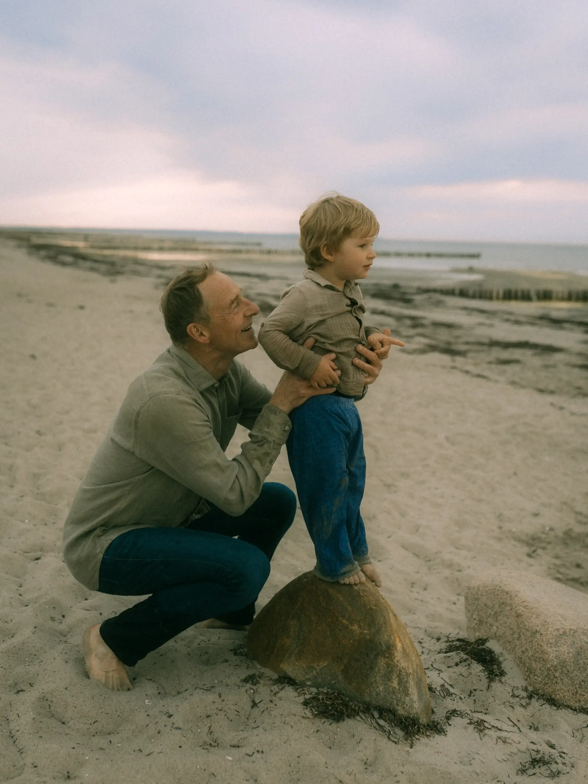 Ein liebevoller Familienfotograf in Schwerin hält einen kleinen Jungen fest, der barfuß auf einem Stein am Strand steht und in die Ferne blickt. Der Vater trägt ein beiges Hemd und Jeans, während sie gemeinsam einen entspannten Moment an der Küste genießen.