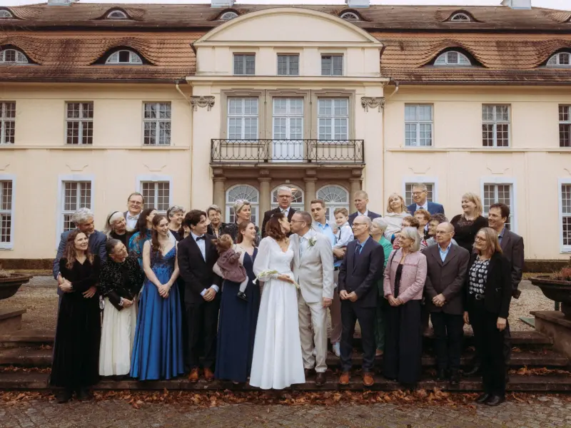 Ein Brautpaar küsst sich vor einem historischen Schloss, umgeben von einer großen Hochzeitsgesellschaft in festlicher Kleidung. Die Aufnahme zeigt die herzliche Stimmung einer Feier, professionell festgehalten von einem Hochzeitsfotograf in Schwerin und MV.