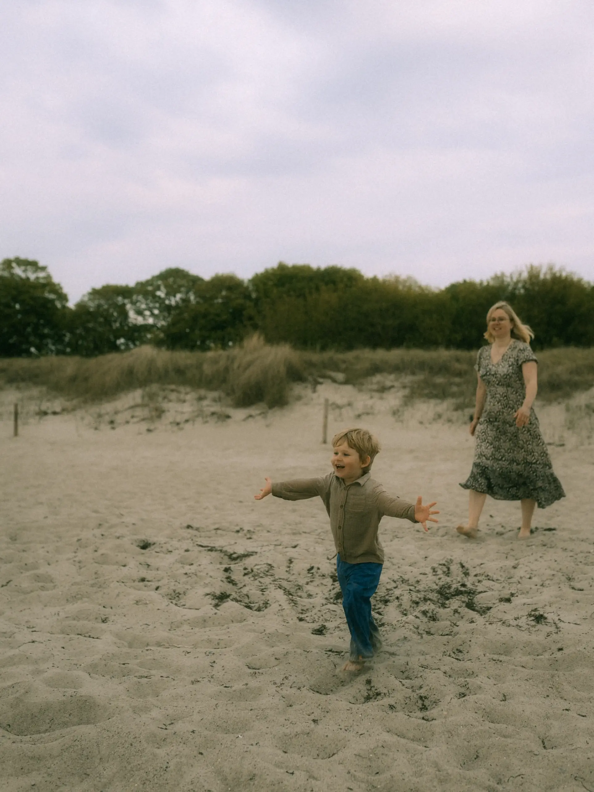 Ein kleiner Junge mit ausgebreiteten Armen läuft freudig über einen Sandstrand, während seine Mutter lächelnd im Hintergrund folgt. Die natürliche Szene am Meer fängt einen glücklichen Moment ein, perfekt festgehalten von einem Fotograf für Familien in Schwerin.