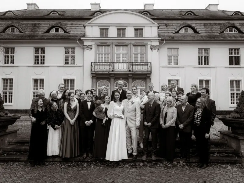 Eine große Hochzeitsgesellschaft posiert vor einem historischen, weißen Herrenhaus. Das Brautpaar steht im Zentrum, umgeben von Familie und Freunden in festlicher Kleidung. Professionelle Hochzeitsfotografie durch einen Hochzeitsfotograf in Schwerin und MV.