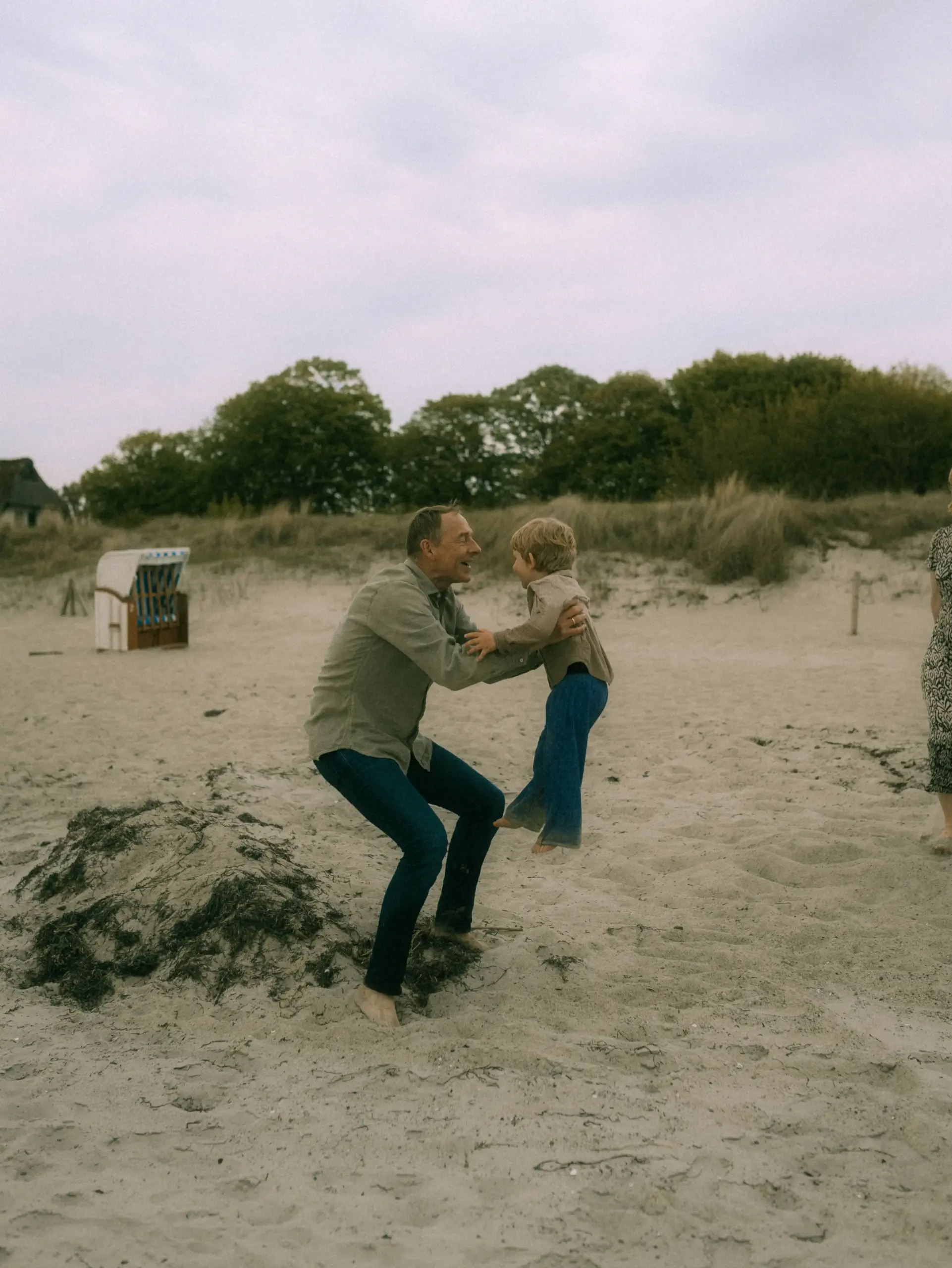 Ein liebevoller Familienfotograf in Schwerin hält einen kleinen Jungen am Strand hoch, während beide sich freudig anlächeln. Die Szene am sandigen Küstenabschnitt mit Dünen im Hintergrund fängt einen herzlichen Moment fest, der typisch für natürliche Familienfotos in Schwerin ist.