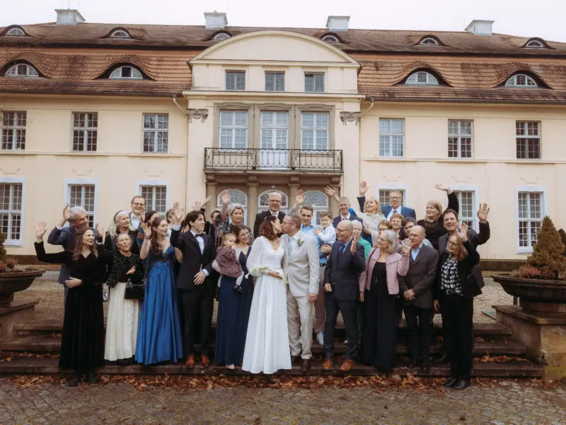 Ein Brautpaar küsst sich vor einer eleganten Schlossfassade, umgeben von einer großen Hochzeitsgesellschaft, die fröhlich winkt. Das Bild wurde von einem Hochzeitsfotograf in Schwerin und MV festgehalten und fängt die festliche Stimmung einer Hochzeit im Freien ein.