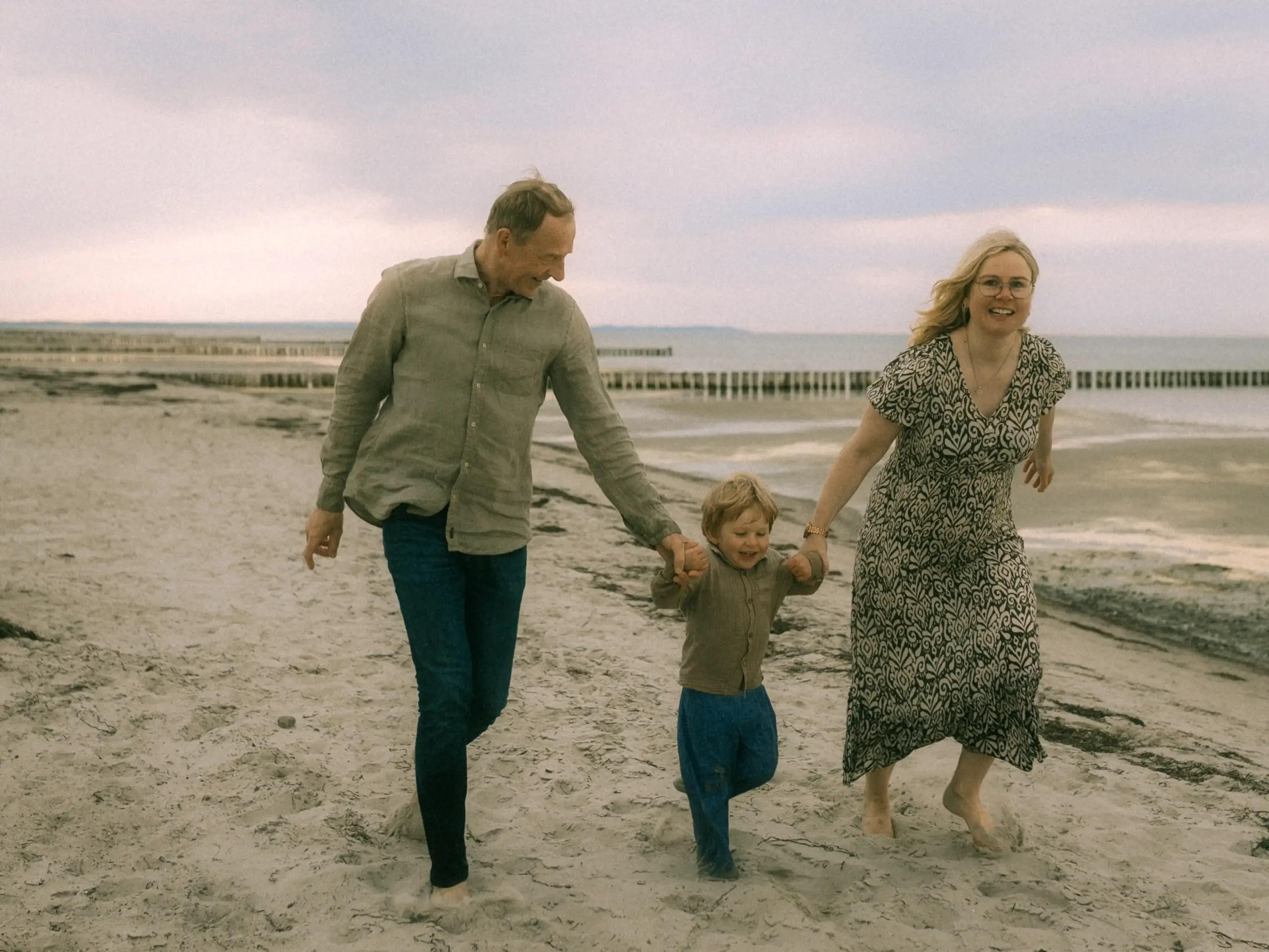 Ein glückliches Paar mit ihrem kleinen Sohn läuft lachend Hand in Hand am Strand entlang. Die Familie genießt einen entspannten Tag am Meer, eingefangen von einem professionellen Familienfotograf in Schwerin, der natürliche Familienfotos in Schwerin kreiert.