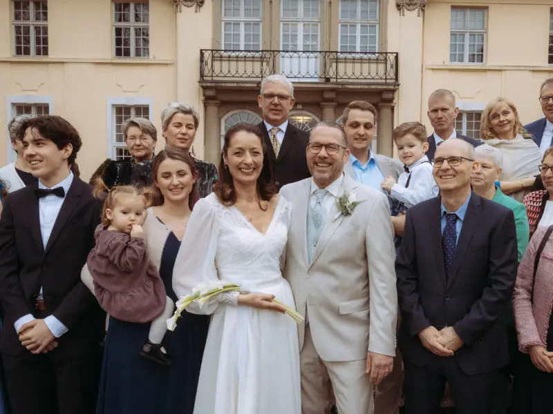 Ein glückliches Brautpaar posiert vor einem historischen Schloss mit Gästen für ein Gruppenfoto, festgehalten von einem Hochzeitsfotograf in Schwerin und MV. Die Braut trägt ein elegantes weißes Kleid, der Bräutigam einen hellen Anzug, umgeben von Familie und Freunden.