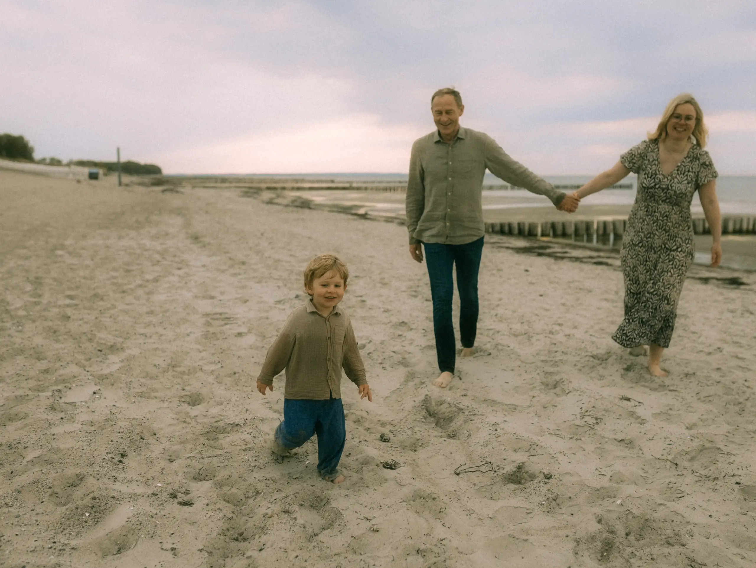 Ein glückliches Paar läuft mit seinem kleinen Sohn barfuß am Strand entlang. Die Familie genießt den gemeinsamen Spaziergang im Sand bei bewölktem Himmel. Professionelle Familienfotos in Schwerin, die eine natürliche und entspannte Stimmung einfangen.