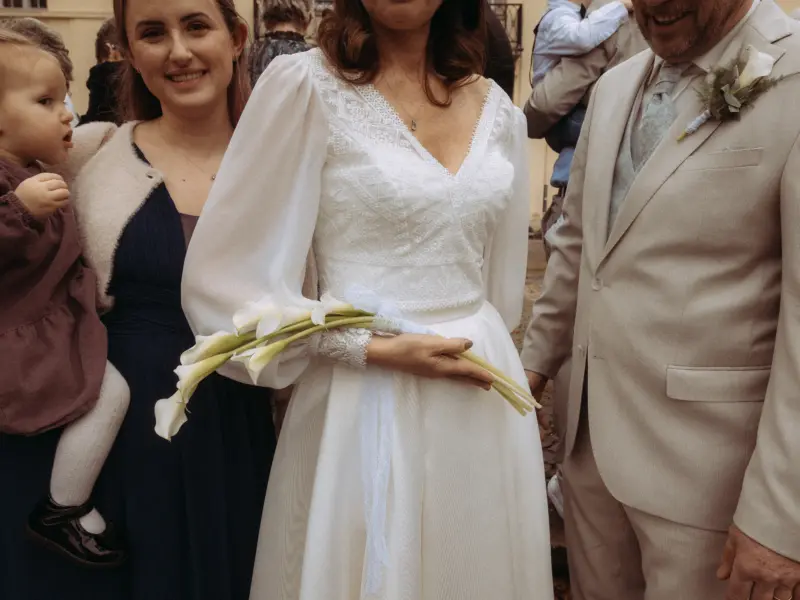 Eine Braut im eleganten weißen Kleid mit Spitzenbesatz und weiten Ärmeln hält einen Brautstrauß aus Calla-Lilien. Neben ihr steht ein Bräutigam im beigen Hochzeitsanzug, während links eine Frau ein Kleinkind auf dem Arm hält. Hochzeitsfotograf in Schwerin und MV.