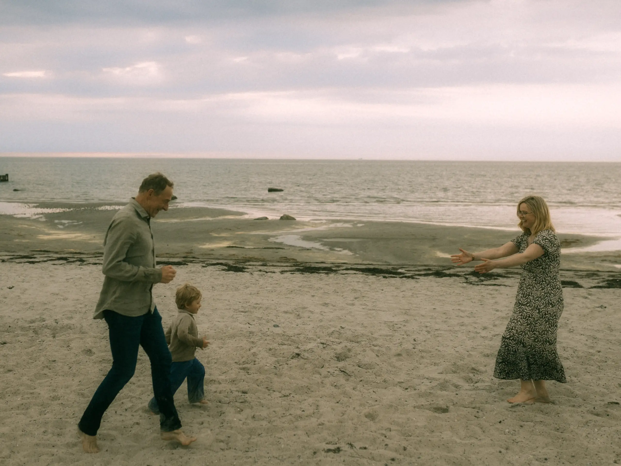 Ein glücklicher Familienmoment am Strand: Ein Vater und sein kleiner Sohn laufen barfuß über den Sand auf die Mutter zu, die mit offenen Armen wartet. Ein natürliches Familienfoto, aufgenommen vom Fotograf für Familien in Schwerin, vor der weiten Kulisse des Meeres an einem bewölkten Tag.