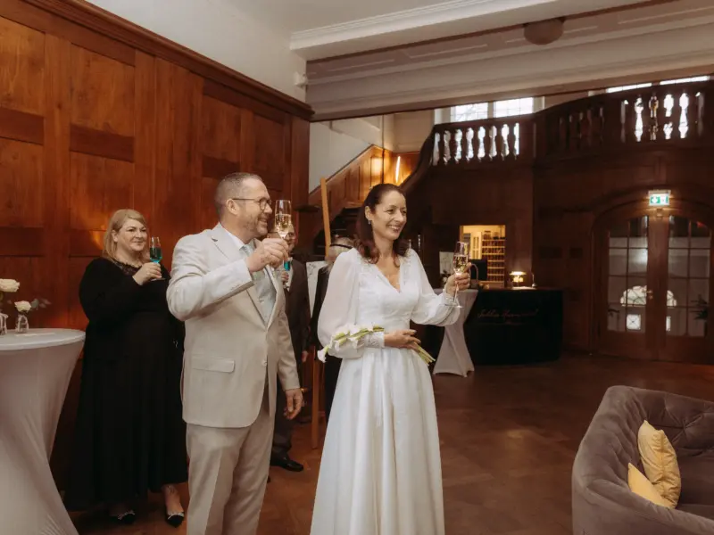 Ein glückliches Brautpaar stößt mit Champagnergläsern in einem eleganten, holzgetäfelten Saal an. Die Braut trägt ein weißes Langarmkleid, der Bräutigam einen hellen Anzug. Im Hintergrund ist eine Treppe zu sehen. Aufnahme von einem Hochzeitsfotograf in Schwerin und MV.