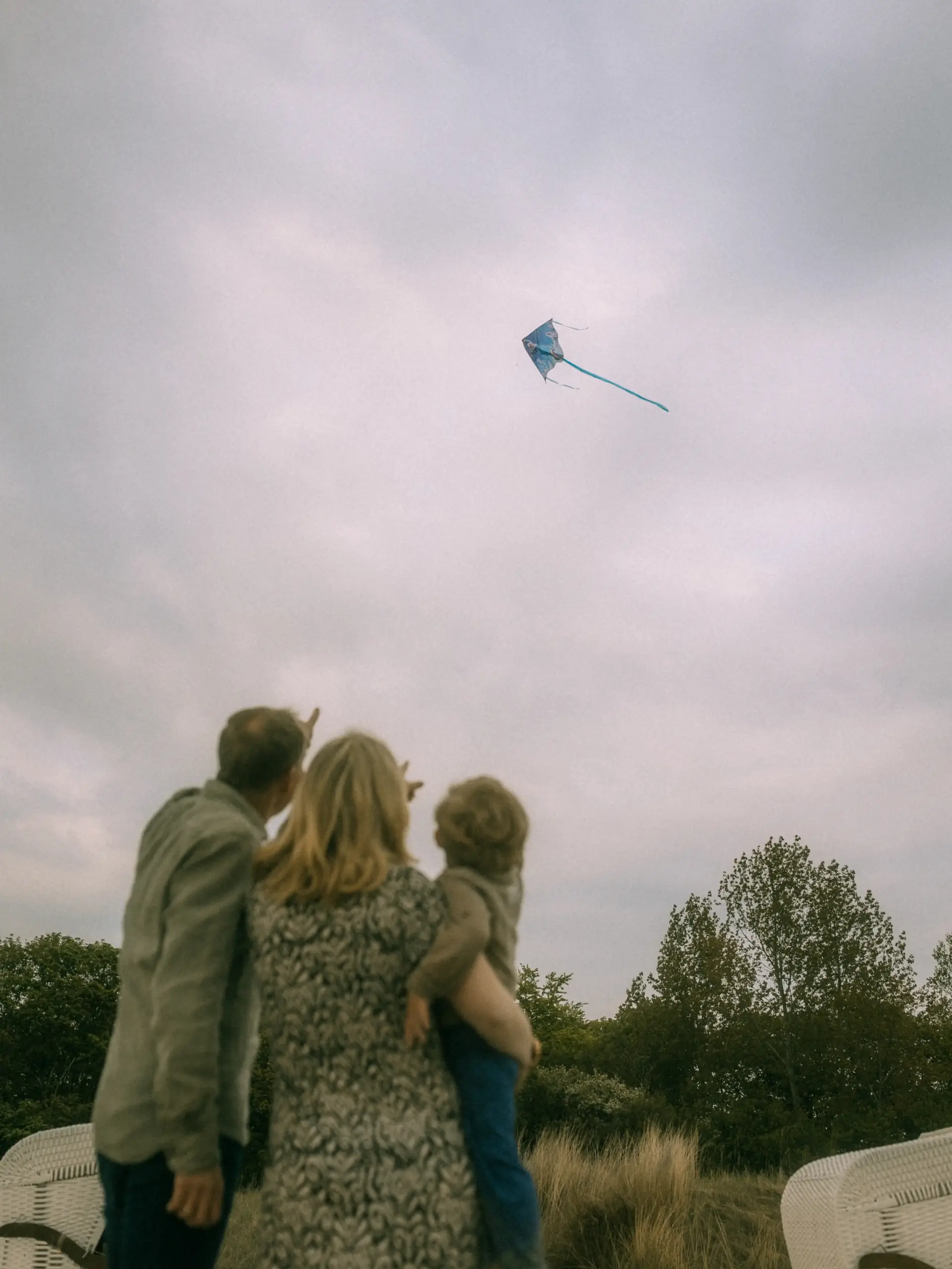 Eine Familie blickt gemeinsam in den bewölkten Himmel, während sie einen blauen Drachen steigen lässt. Der Vater, die Mutter und ihr Kind stehen in einer natürlichen Dünenlandschaft. Authentische Familienfotos in Schwerin vom Familienfotograf in Schwerin.