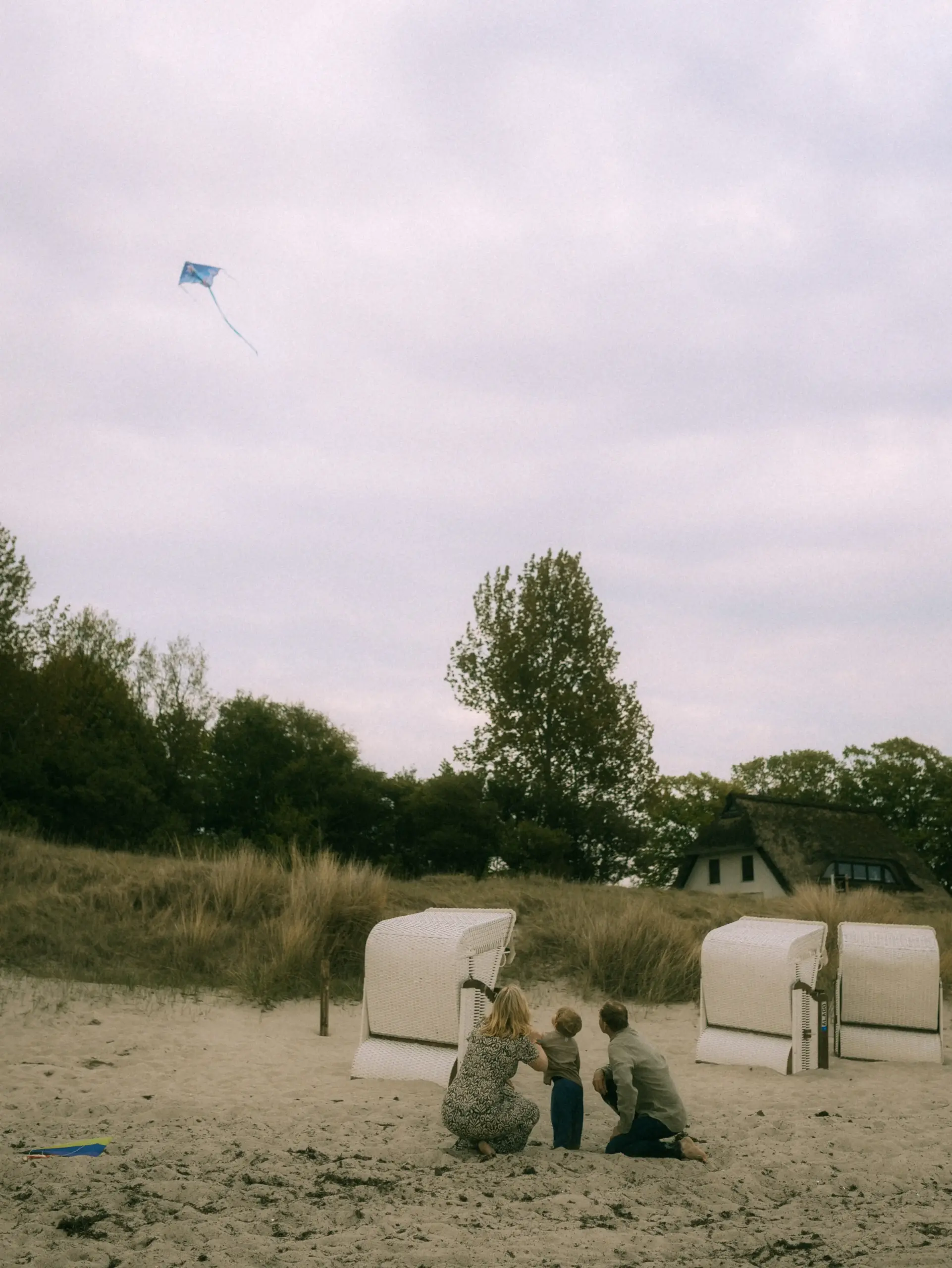 Eine junge Familie lässt gemeinsam einen blauen Drachen am Strand steigen. Die Eltern und ihr Kind knien im Sand vor typischen Strandkörben, während im Hintergrund eine Dünenlandschaft mit einem reetgedeckten Haus zu sehen ist. Professionelle Familienfotos in Schwerin.
