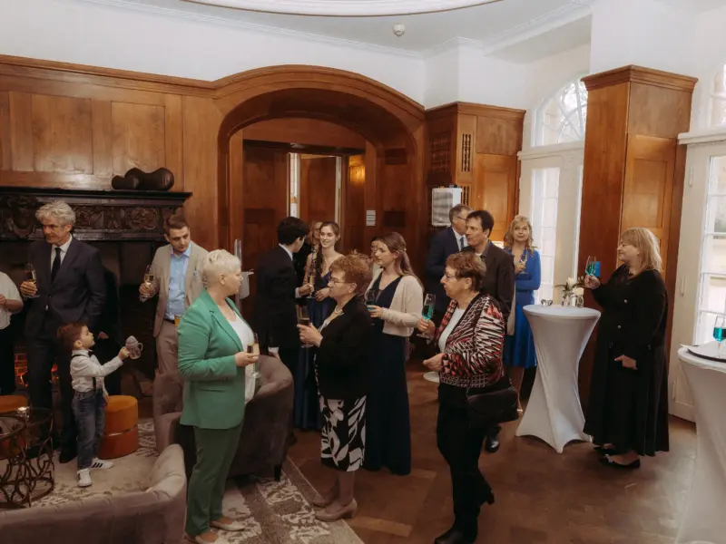 Gäste bei einem eleganten Sektempfang in einem holzgetäfelten Saal, festgehalten von einem Hochzeitsfotograf in Schwerin und MV. Die Gesellschaft unterhält sich bei Getränken, während ein kleiner Junge im Anzug den Raum erkundet, umrahmt von der stilvollen Atmosphäre einer Hochzeitsfeier.