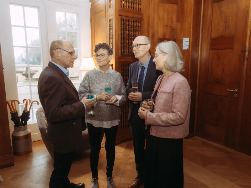 Zwei Paare im eleganten Outfit führen ein angeregtes Gespräch bei einem Empfang in einem stilvollen Raum mit Holzvertäfelung. Sie halten Sekt- und Cocktailgläser in der Hand, während im Hintergrund eine Fensterfront den Blick nach draußen freigibt. Fotograf für Familien in Schwerin.