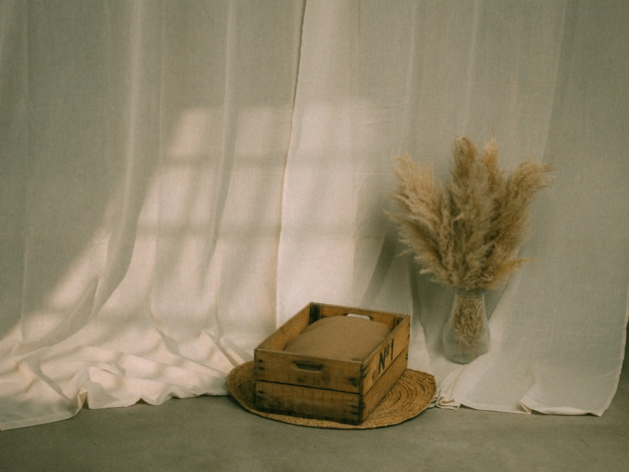 Wooden crate with sand on straw mat, pampas grass in vase, draped fabric