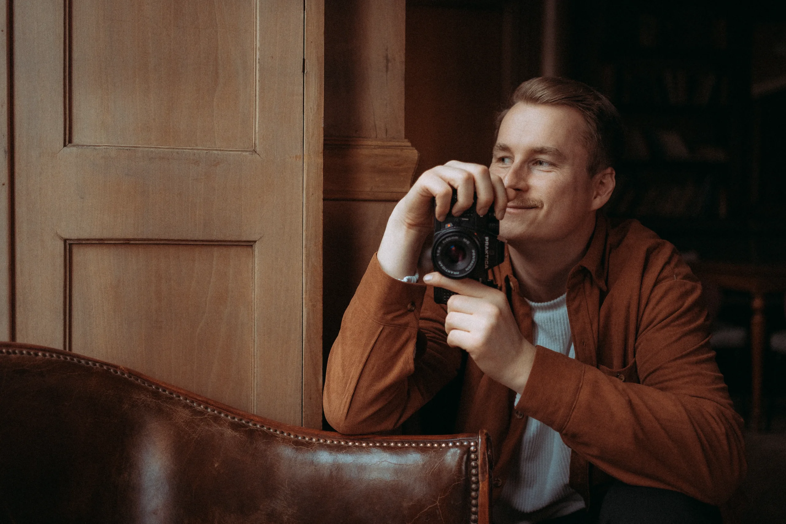 Ein lächelnder Fotograf hält eine analoge Praktica-Kamera in der Hand und schaut nachdenklich zur Seite. Er trägt ein rostbraunes Hemd und sitzt in einem gemütlichen Raum mit Ledersessel. Als Hochzeitsfotograf in Schwerin und MV fängt er besondere Momente mit Leidenschaft ein. Ein lächelnder Fotograf hält eine analoge Praktica-Kamera in der Hand und schaut nachdenklich zur Seite. Er trägt ein rostbraunes Hemd und sitzt in einem gemütlichen Raum mit Ledersessel. Als Hochzeitsfotograf in Schwerin und MV fängt er besondere Momente mit Leidenschaft ein.