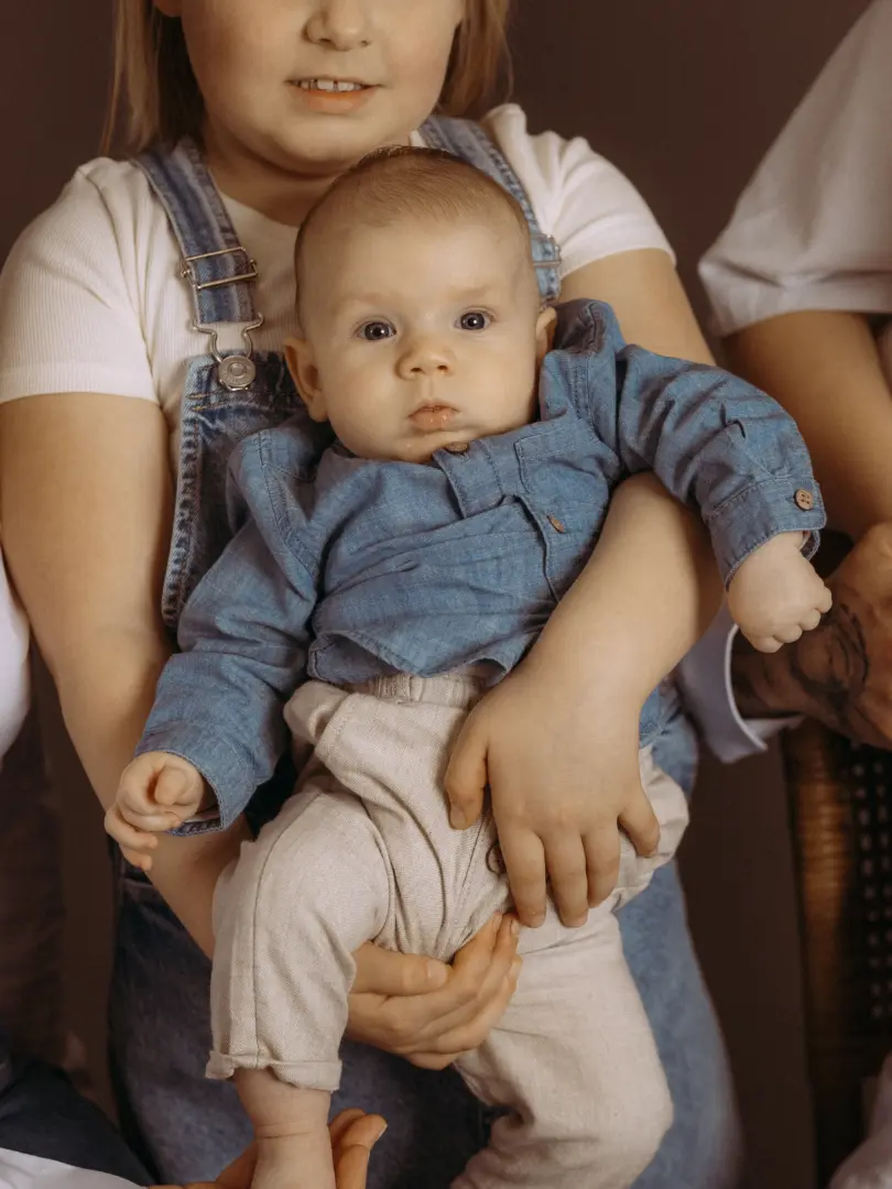 Ein Baby mit wachem Blick trägt ein blaues Jeanshemd und eine helle Hose, während es liebevoll von einer Person in einer blauen Latzhose gehalten wird. Das stimmungsvolle Porträt entstand als professionelles Familienfoto im Fotostudio in Schwerin.