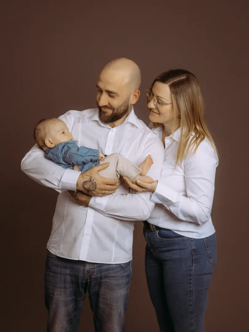 Ein glückliches Paar hält liebevoll sein Baby in einem Fotostudio in Schwerin. Die Eltern tragen weiße Hemden und Jeans vor einem schlichten braunen Hintergrund, während sie ihren kleinen Sohn betrachten. Ein professionelles Familienfoto vom Fotograf für Familien in Schwerin.
