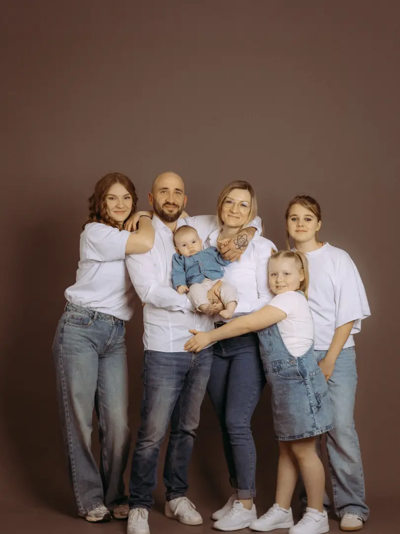Eine glückliche sechsköpfige Familie posiert gemeinsam vor einem schlichten braunen Hintergrund in einem Fotostudio in Schwerin. Eltern, drei Töchter und ein Baby tragen weiße Oberteile und Jeans für ein zeitloses Familienfoto vom professionellen Familienfotograf in Schwerin.