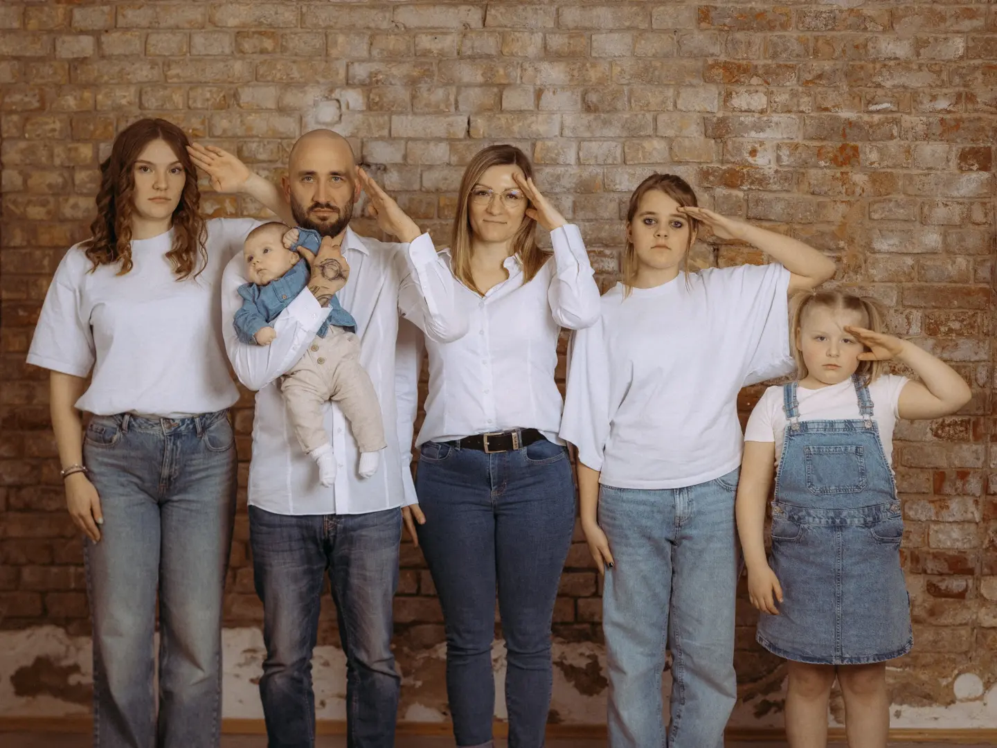 Eine Familie steht vor einer Ziegelwand und salutiert in die Kamera. Der Vater hält ein Baby im Arm. Die Familienmitglieder tragen weiße Oberteile und Jeans. Dieses Bild entstand bei einem Shooting für Familienfotos in Schwerin im Fotostudio in Schwerin.