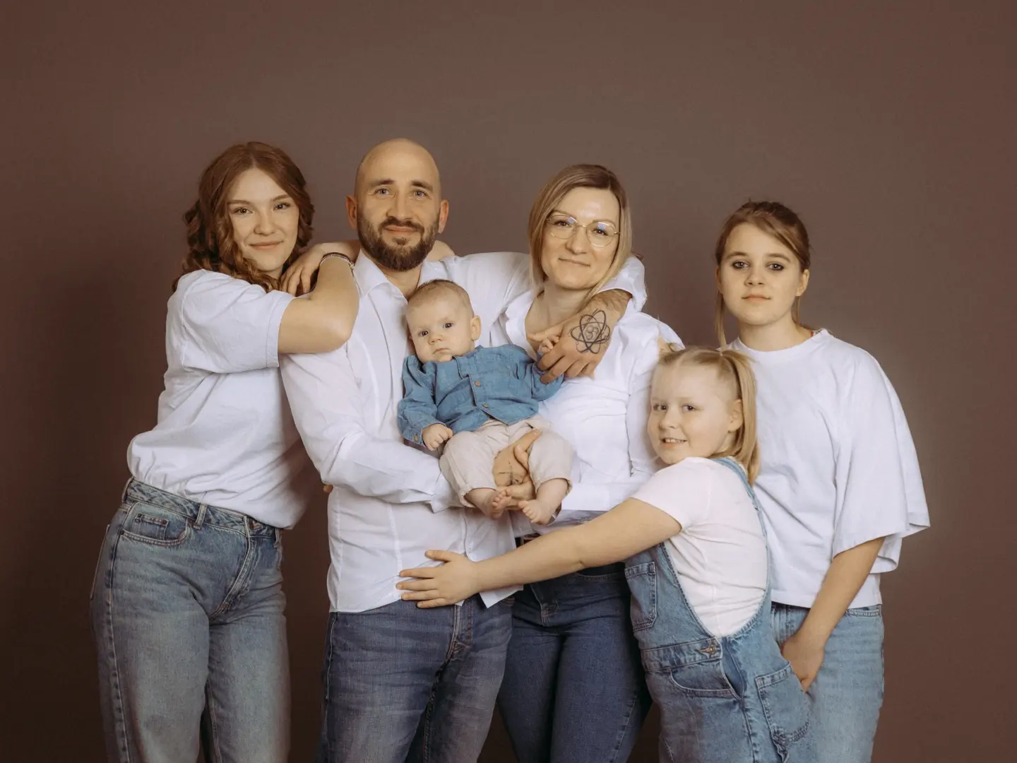Ein Familienfotograf in Schwerin hat dieses Porträt einer sechsköpfigen Familie im Fotostudio in Schwerin aufgenommen. Die Eltern tragen weiße Hemden und Jeans, während sie ihre vier Kinder vor einem schlichten braunen Hintergrund halten und umarmen, was eine herzliche Atmosphäre schafft.
