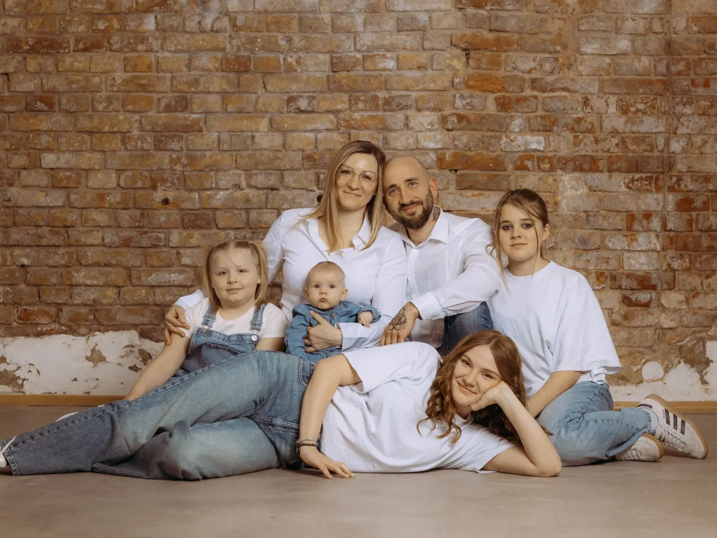 Eine glückliche Familie posiert für ein professionelles Foto in einem Fotostudio in Schwerin. Vor einer rustikalen Backsteinwand sitzen Eltern mit ihren vier Kindern, darunter ein Baby, in harmonischen weißen Outfits und Jeans, was für ein zeitloses Familienfoto sorgt.