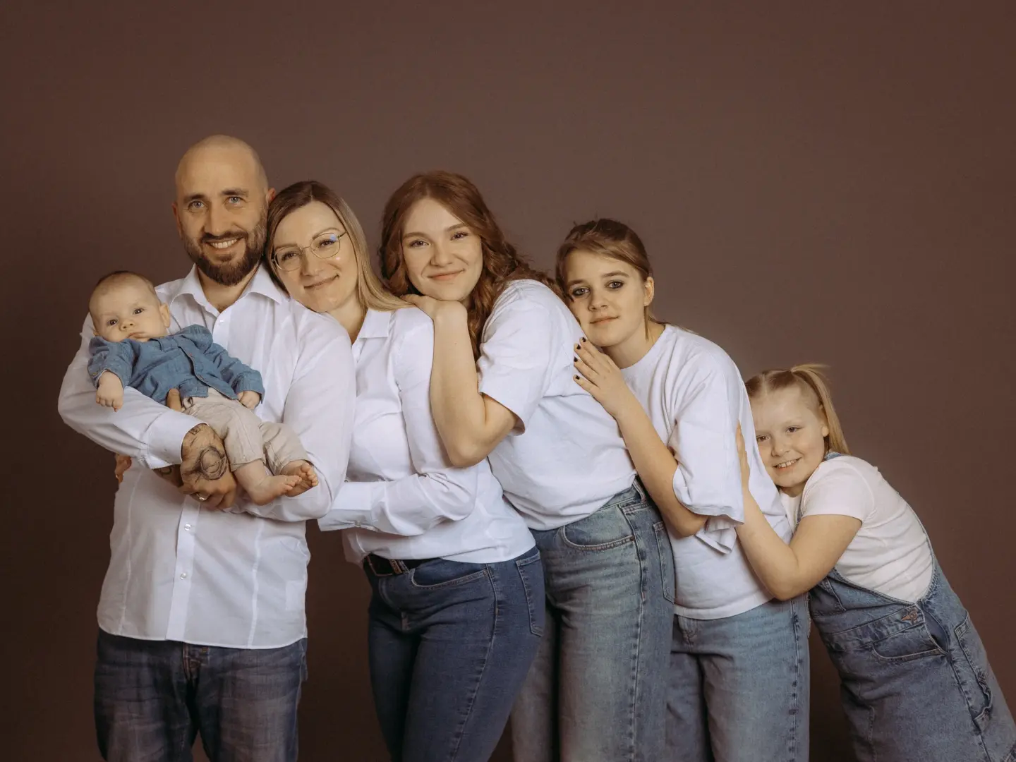 Eine glückliche Familie posiert für ein professionelles Foto im Fotostudio in Schwerin. Ein Vater hält sein Baby im Arm, neben ihm stehen die Mutter und ihre drei Töchter in einer Reihe, alle tragen weiße Oberteile und Jeans vor einem schlichten, braunen Hintergrund.