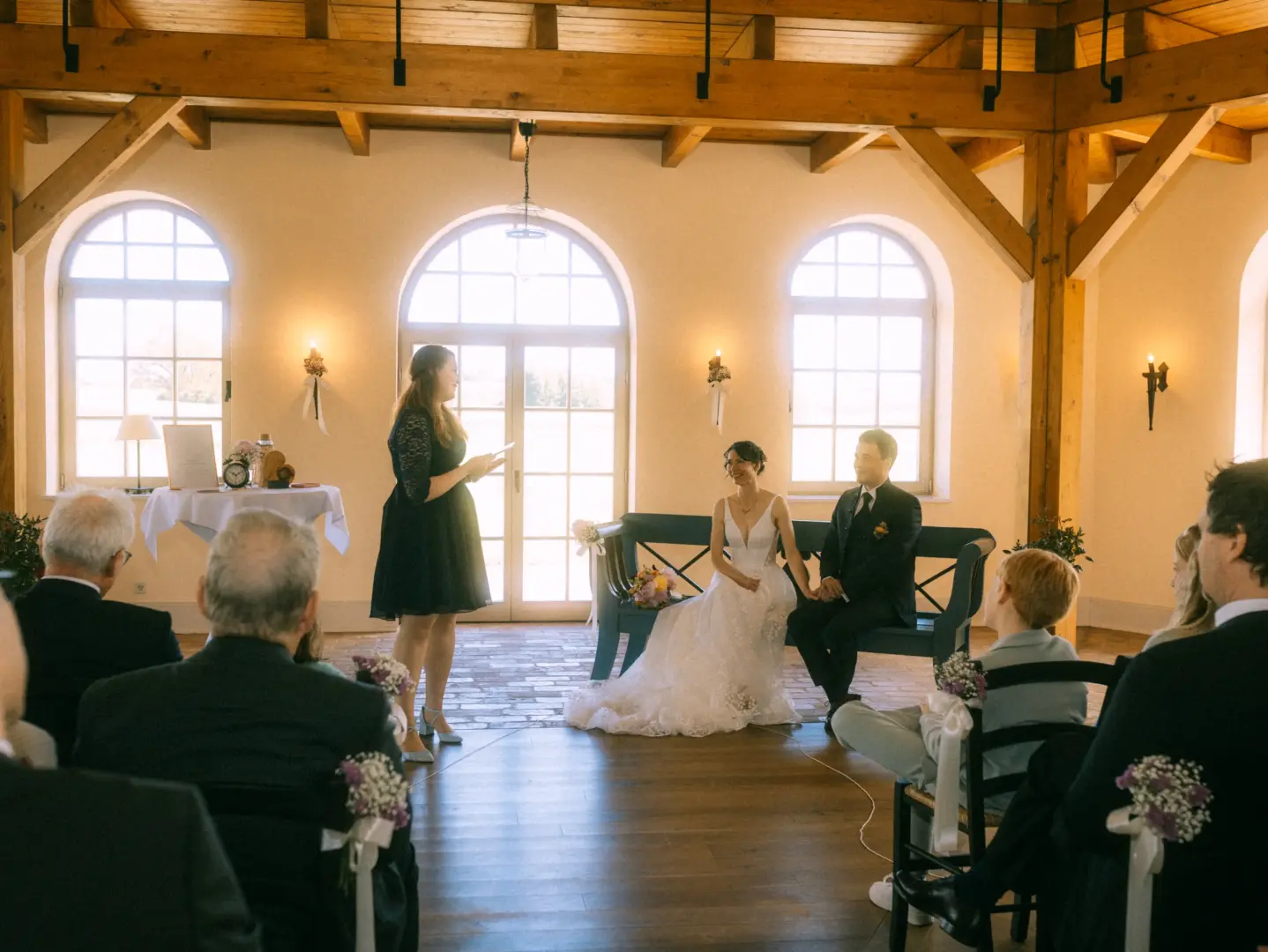 Eine Braut im weißen Spitzenkleid und ihr Bräutigam im dunklen Anzug sitzen während ihrer Trauung auf einer blauen Bank, während eine Rednerin spricht. Hochzeitsfotograf in Schwerin und MV, festgehalten in einem hellen, rustikalen Saal mit großen Fenstern und Holzbalken.