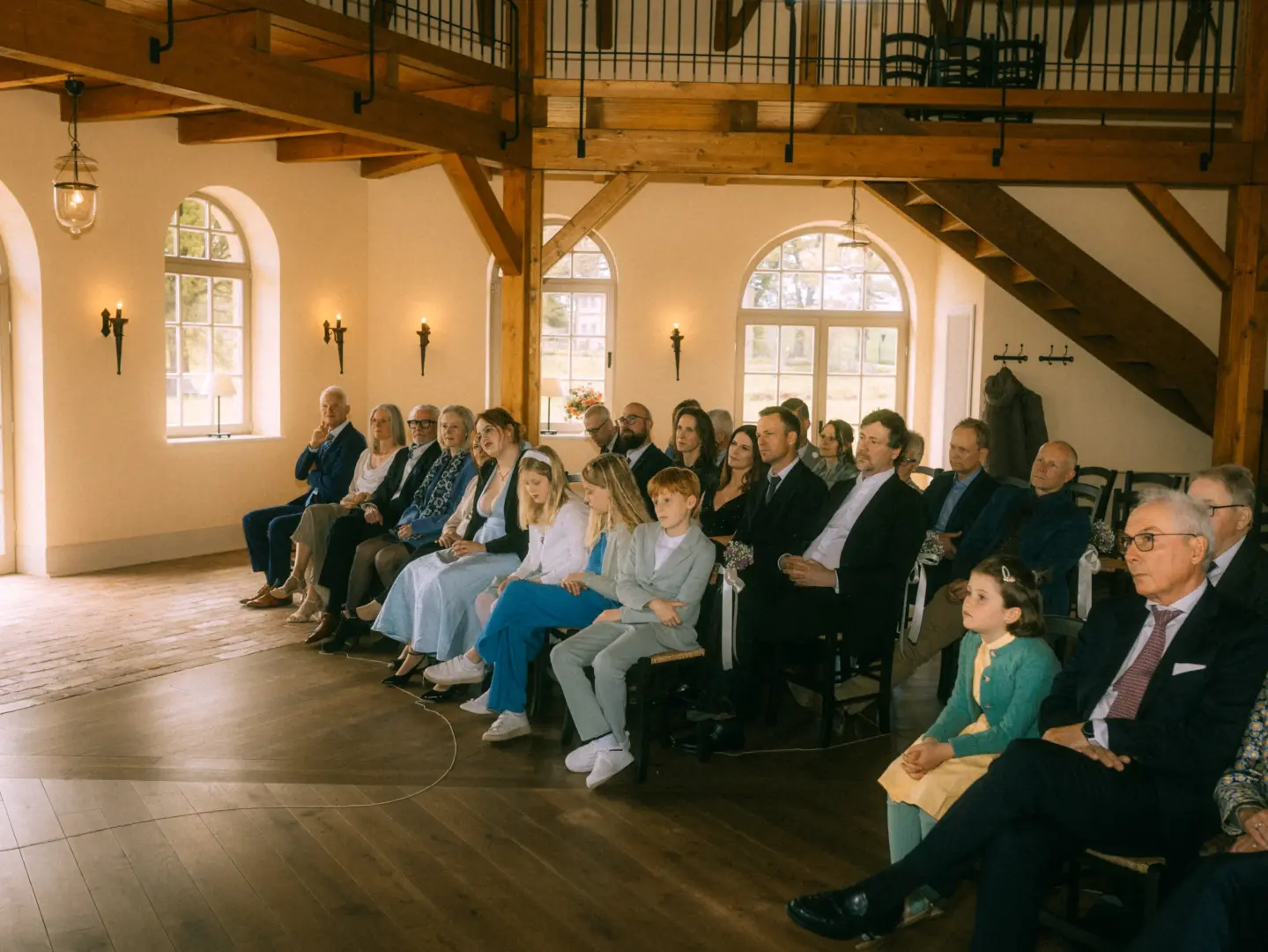 Gäste sitzen in einer eleganten, lichtdurchfluteten Scheune bei einer Hochzeitszeremonie. Die Hochzeitsgesellschaft, bestehend aus Familien und Freunden, verfolgt aufmerksam das Geschehen. Professionelle Aufnahme von einem Hochzeitsfotograf in Schwerin und MV in einem rustikalen Ambiente.