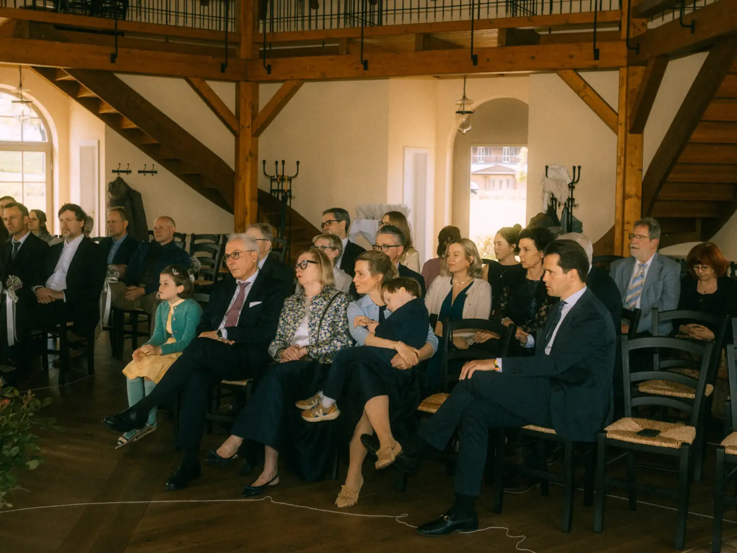 Gäste sitzen während einer Hochzeitszeremonie in einem hellen, rustikalen Saal mit Holzbalken. Das Publikum verfolgt aufmerksam das Geschehen, während ein Kind auf dem Schoß seiner Mutter schläft. Hochzeitsfotograf in Schwerin und MV.