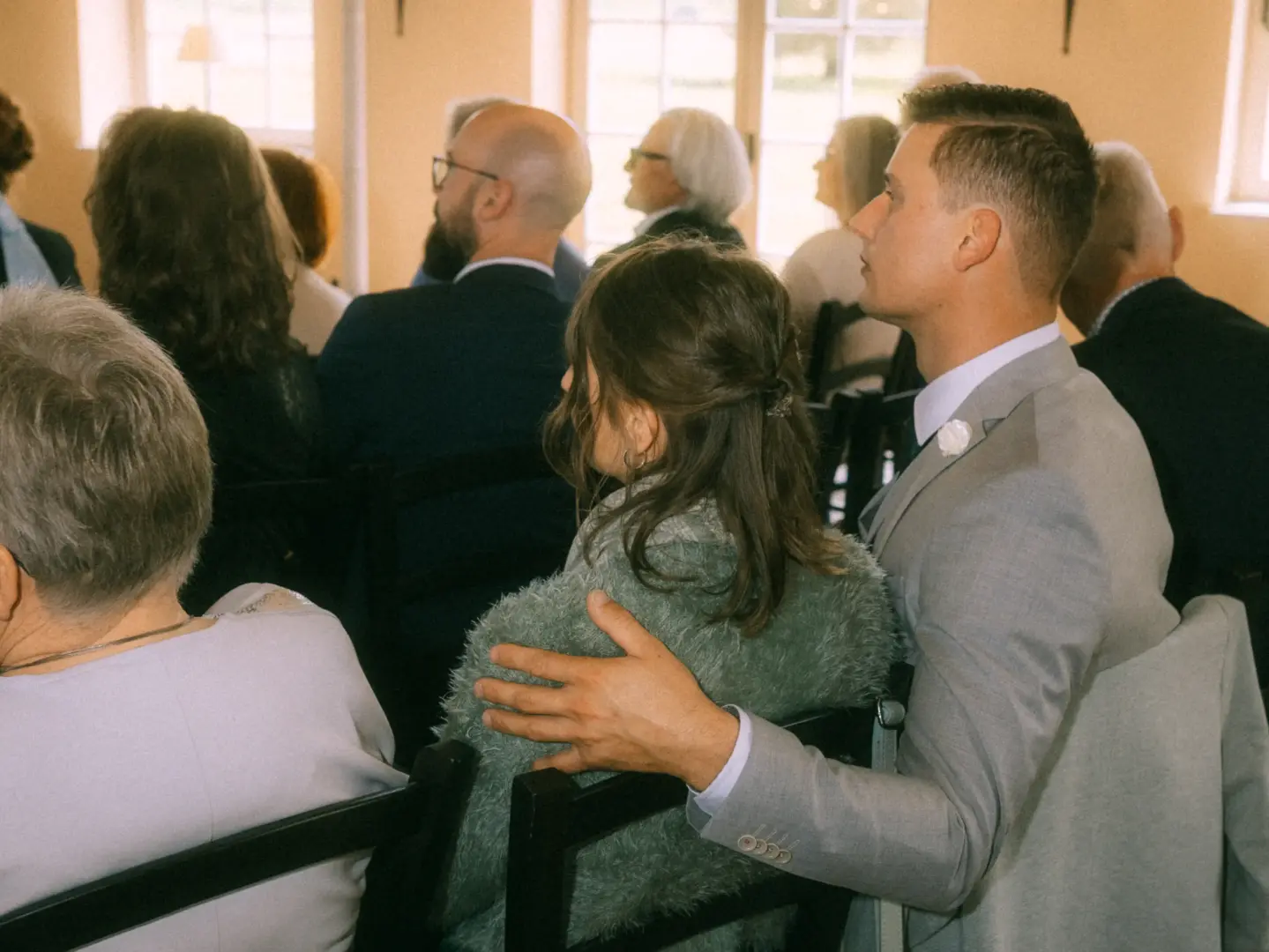 Ein junges Paar sitzt nebeneinander in der ersten Reihe einer Hochzeitszeremonie. Der Mann im hellgrauen Anzug legt liebevoll seinen Arm um die Frau in der flauschigen grünen Strickjacke. Hochzeitsfotograf in Schwerin und MV.