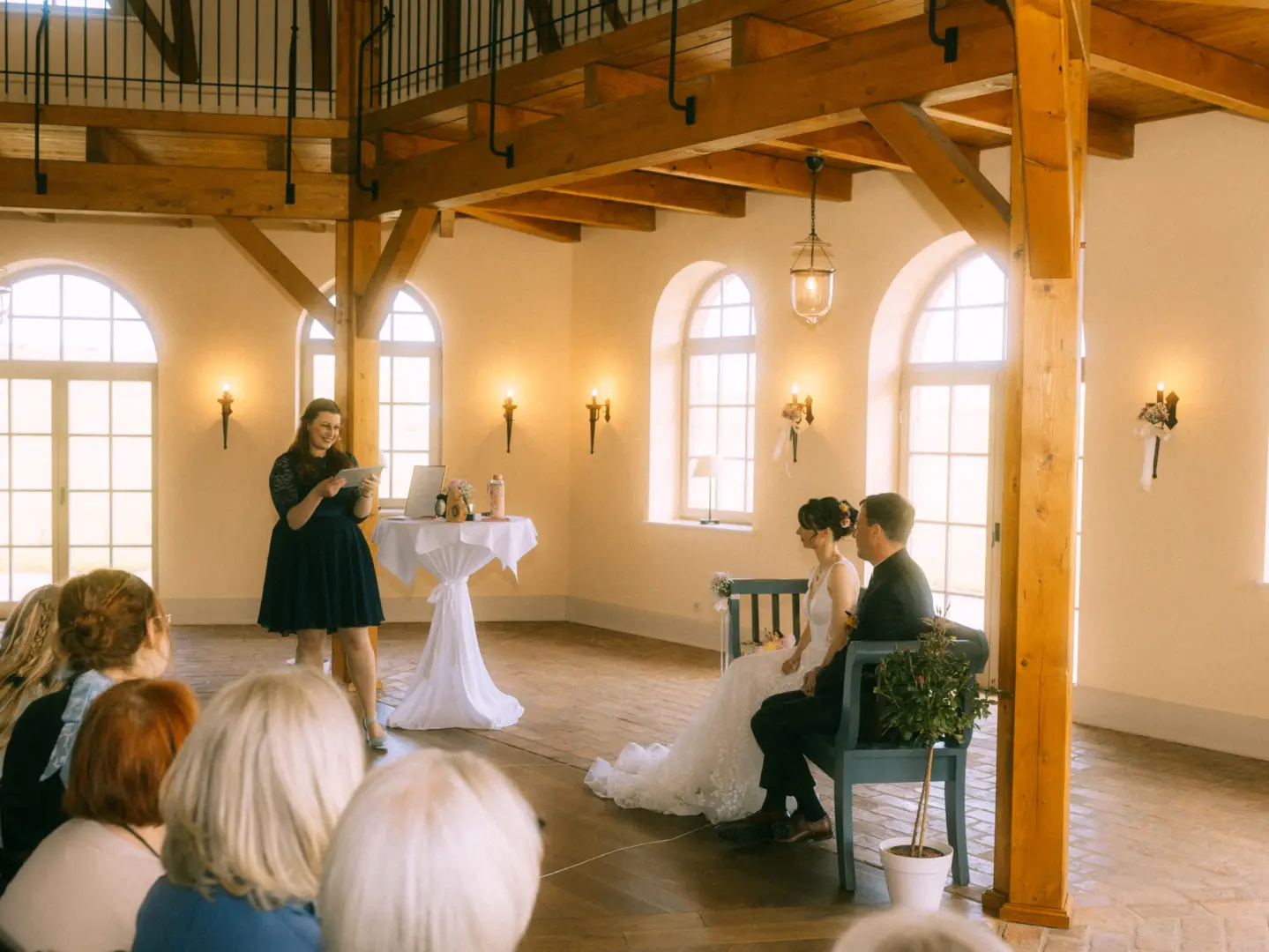 Eine Braut und ein Bräutigam sitzen während ihrer Trauung auf einer blauen Bank in einem lichtdurchfluteten, rustikalen Saal, während eine Rednerin vor ihnen spricht – festgehalten von Ihrem Hochzeitsfotograf in Schwerin und MV.