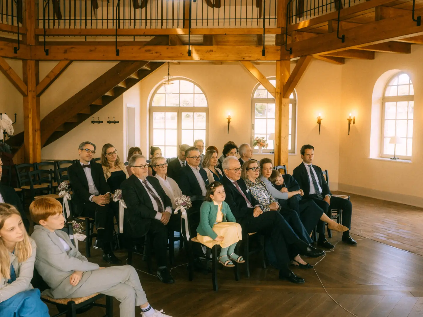 Gäste sitzen in einem eleganten, rustikalen Saal mit Holzbalken bei einer Hochzeitszeremonie. Die festliche Atmosphäre wird von einem Hochzeitsfotograf in Schwerin und MV festgehalten, während die aufmerksamen Zuschauer gespannt nach vorne blicken.