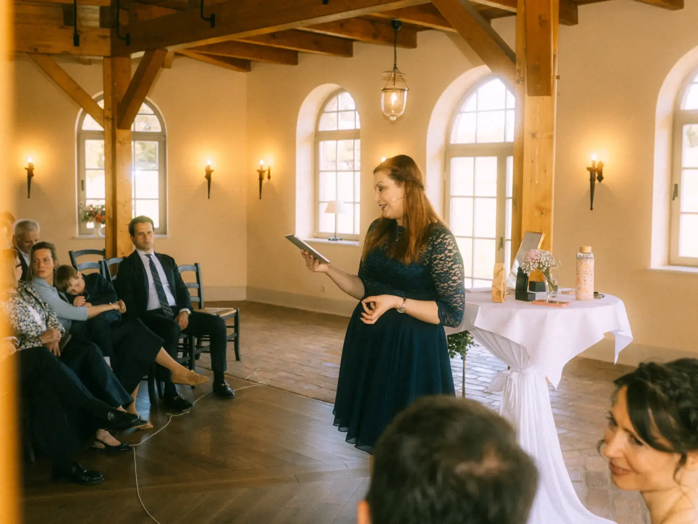 Eine Frau in einem dunkelblauen Spitzenkleid hält eine Rede während einer Trauung in einem hellen, rustikalen Saal mit Holzbalken. Gäste sitzen aufmerksam auf Stühlen, während ein Hochzeitsfotograf in Schwerin und MV diesen emotionalen Moment festhält.